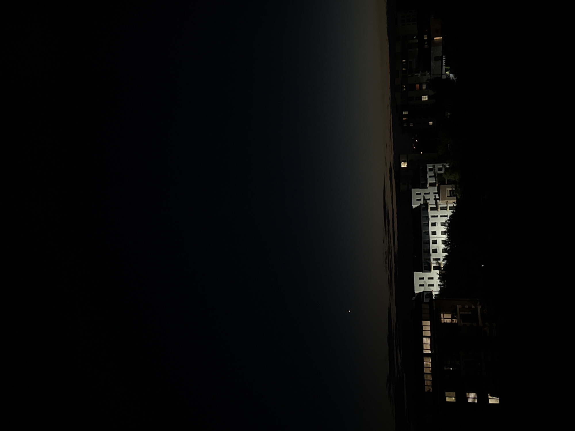 This is a nighttime photograph taken from a high vantage point overlooking a city. The sky is dark with a subtle gradient of light near the horizon, suggesting either dawn or dusk. A small, bright celestial body, possibly Venus, is visible in the upper left quadrant of the sky.
Below the sky, a cityscape emerges from the darkness. Several buildings are illuminated, with windows showing warm light, indicating occupancy. In the foreground, a modern, multi-story building with a white facade and many windows is prominently featured. Its architectural style suggests residential or office use. To its right, another building, also illuminated, appears to be part of the same complex or a neighboring structure. Further into the background, more buildings are visible, some with scattered lights, contributing to the urban landscape. The lower portion of the image is almost entirely black, obscuring the ground level and any potential details there. Trees and foliage are subtly silhouetted against the lit buildings, adding a touch of nature to the urban scene. The overall impression is one of a peaceful, illuminated urban environment at night.