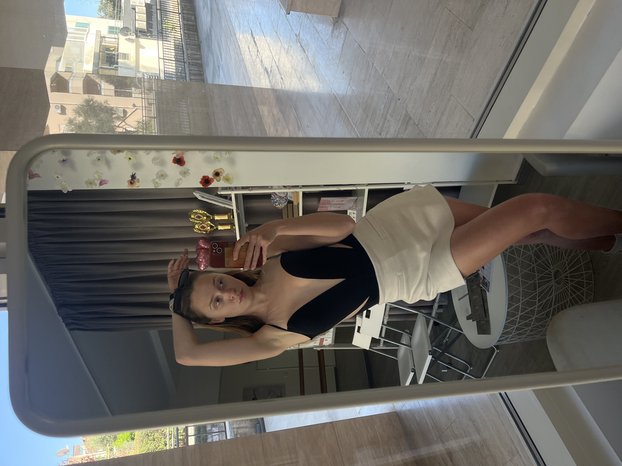 A fair-skinned woman with blonde hair is taking a selfie in a mirror. She is wearing a black one-piece swimsuit with a large cutout on the left side of her torso, paired with a white skirt. Her left arm is raised with sunglasses in hand, while her right hand holds a mobile phone to take the picture.
The mirror is reflecting the scene within a room with a shelf decorated with artificial flowers and decorative objects. A white round table and chairs are visible near the woman.
The reflection in the mirror shows an exterior scene with buildings and trees, indicating an urban setting. Based on architectural style and general appearance, the location is likely in Budva, Montenegro.