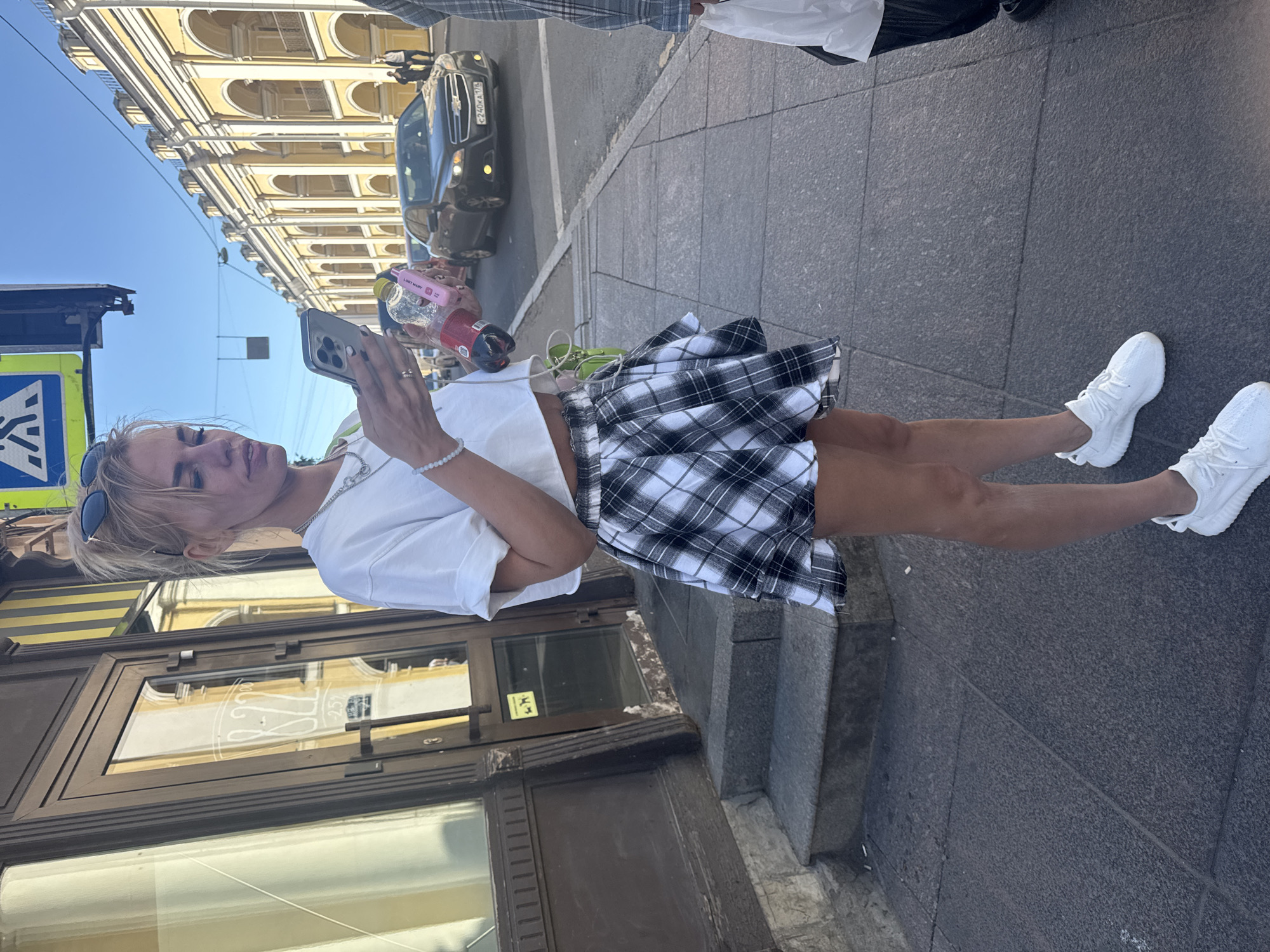 In this vertical, medium shot, a young woman with blonde hair and sunglasses is captured from the mid-thigh up, holding a smartphone and a drink. She is standing on a cobblestone sidewalk next to a dark gray building with large glass windows and an ornate yellow building with arched windows in the background. She is wearing a white t-shirt, a black and white plaid skirt, and white sneakers. A silver necklace and a beaded bracelet adorn her left wrist. The woman appears to be looking at her phone and holding a drink in her right hand. A dark colored car is parked on the street behind her. The scene is outdoors under a clear blue sky, suggesting it is daytime with pleasant weather. A blue and yellow pedestrian crossing sign is visible in the lower left corner of the image. The reflections in the building windows suggest the presence of signage, but no specific text is clearly legible. The overall impression is that of a casual street scene, possibly in a city environment given the architecture and the presence of a car.