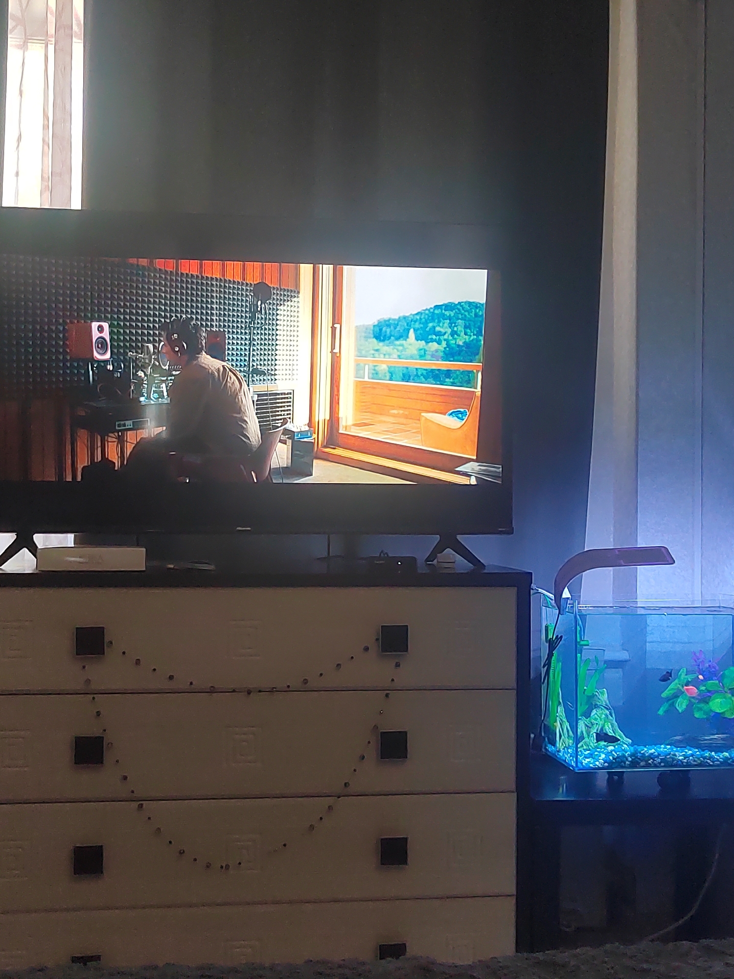 The image shows a television screen displaying a scene of a person in a recording studio. The studio has soundproofing panels on the walls and a microphone in front of the person. The television is placed on top of a dresser with several drawers. To the right of the dresser, there is a small fish tank with a blue light illuminating the water. The room appears to be a domestic setting, possibly a living room or bedroom. The location of Biysk, Russia, is not discernible from the visual information provided in the image.