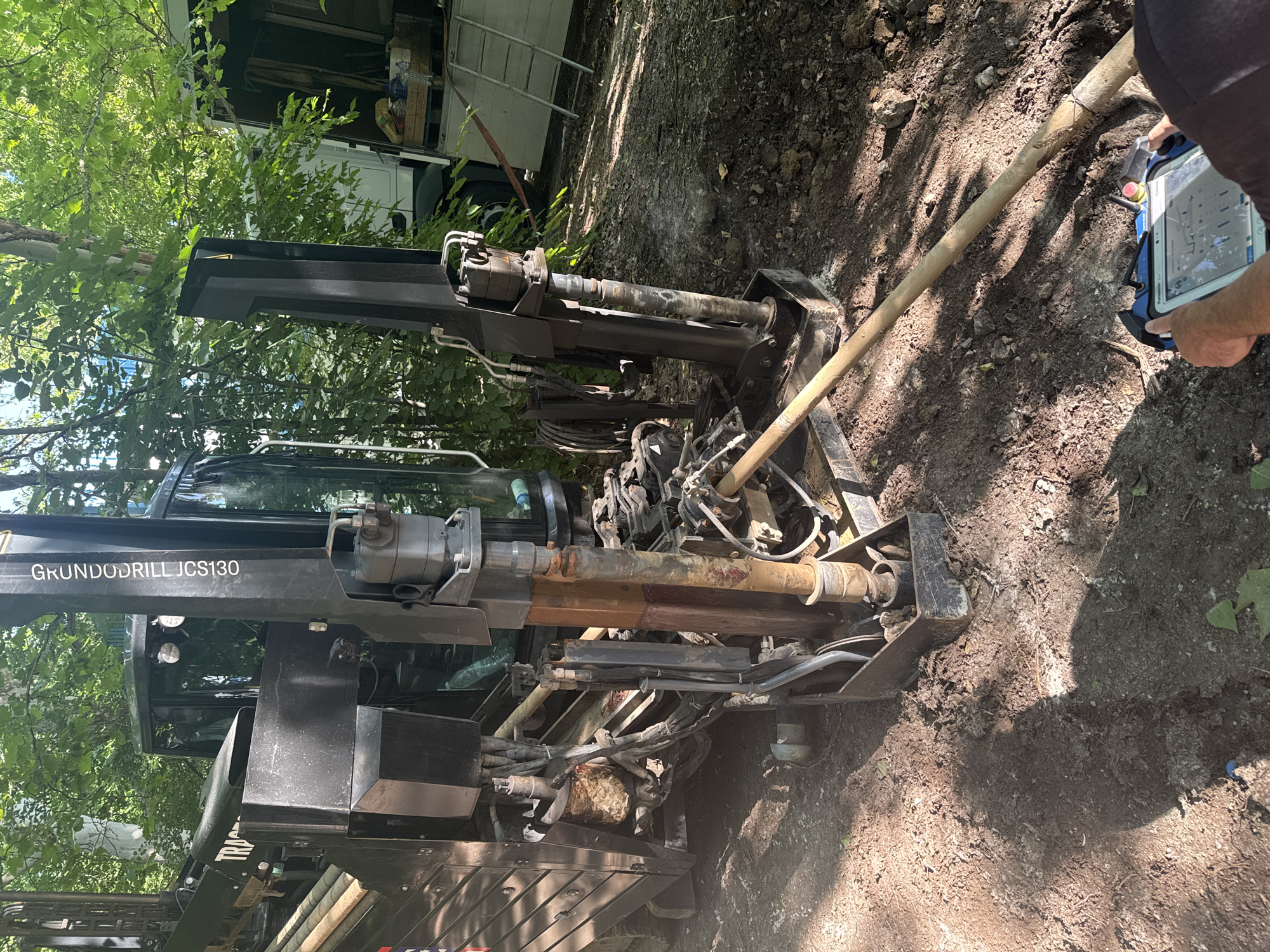 The image shows a horizontal directional drilling rig operating in a wooded area, likely on the outskirts of Pleven, Bulgaria. The rig, identified by the text "GRUNDODRILL JCS130" on its side, is a large piece of machinery with various hydraulic hoses and mechanical components. A long, wooden rod extends from the rig into the ground, suggesting the drilling process is underway. In the foreground, a person's hands are visible holding a remote control device with a screen, indicating they are operating the drill. The environment is characterized by dirt ground, fallen leaves, and dense foliage from trees overhead, with a partial view of a truck in the background. The overall scene depicts an outdoor construction or infrastructure project in progress.