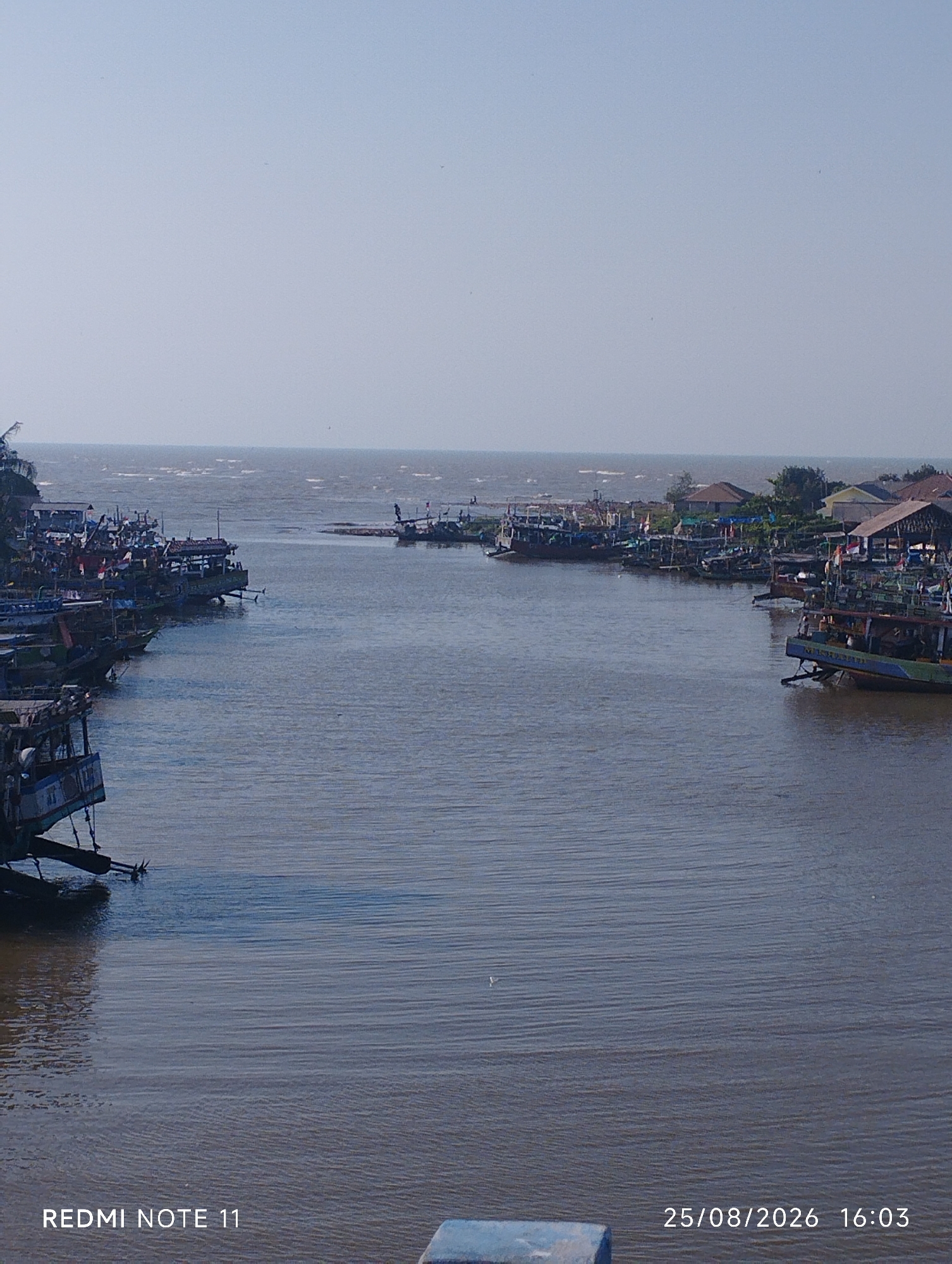 The image depicts a waterfront scene likely in Karawang, Indonesia, with numerous fishing boats moored in a calm river or harbor. The boats are of various sizes and designs, many appearing to be traditional wooden fishing vessels. Some have cabins and masts, while others are more open. They are lined up along the left bank and also visible in the distance on the right bank. The water is a murky brown, reflecting the overcast or hazy sky. On the right bank, buildings with pitched roofs are visible, suggesting a settlement or fishing village. The sky is a pale, washed-out blue, indicating either a hazy day or early morning/late afternoon light. The overall impression is one of a working port or a quiet harbor, with the boats appearing to be at rest. The bottom of the image contains a timestamp indicating "25/08/2026 16:03" and the text "REDMI NOTE 11".