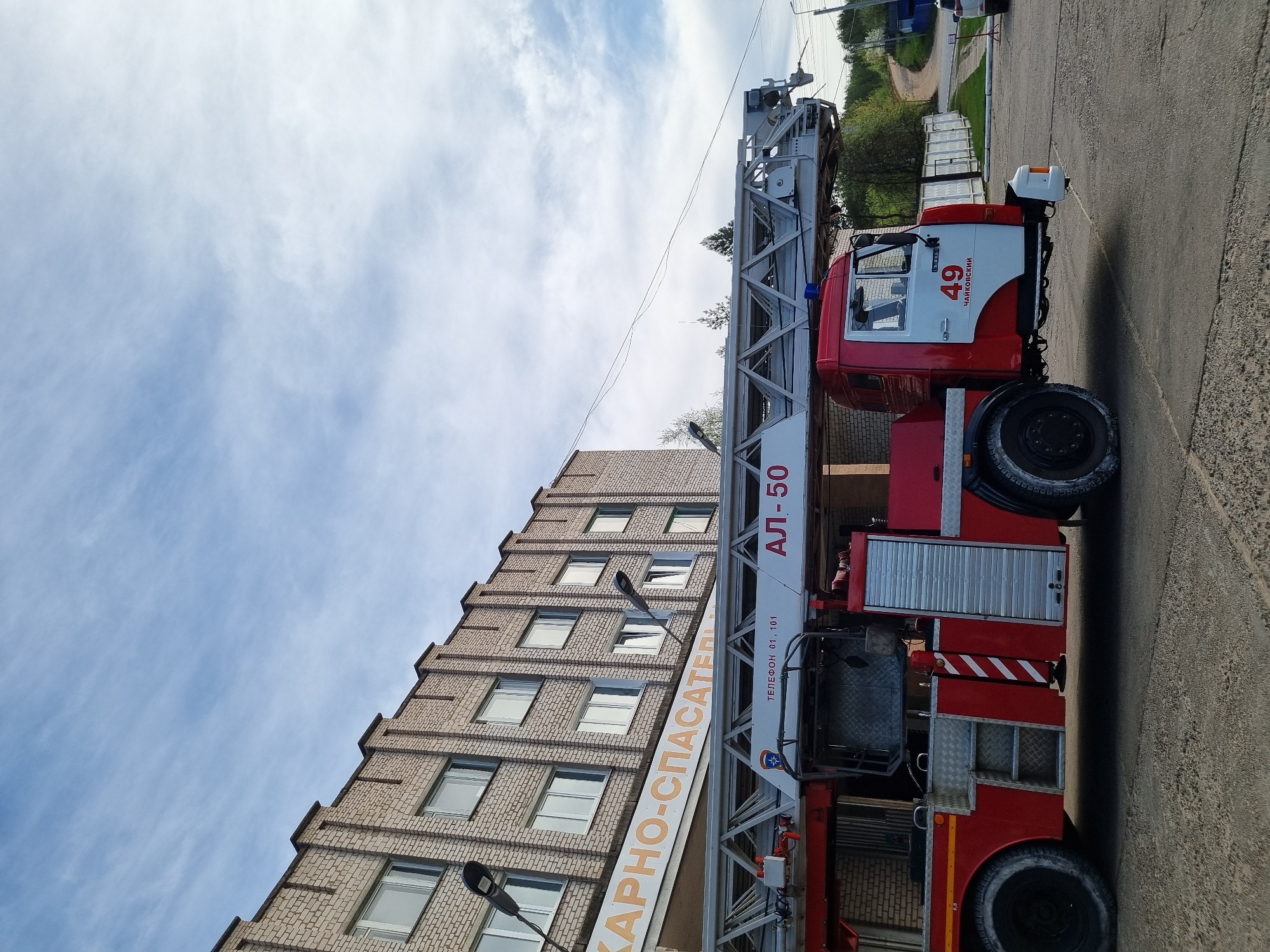 A red and white fire truck with the number 49 and the words "Чайковский" (Chaikovsky) is parked on a paved surface. The fire truck's ladder extends upwards towards a brick building with multiple windows. A sign with the words "КАРНО-СПАСАТЕЛЬ" (KARNO-SAVIOR) is visible on the building. The sky is blue with some clouds. Streetlights are also visible near the building.
