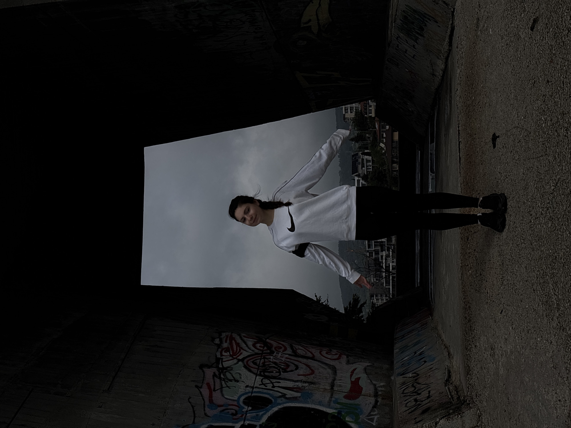 A young woman is standing in a concrete structure in Varna, Bulgaria. The structure appears to be some type of underpass or tunnel. The woman is wearing a white long-sleeved shirt with a Nike logo and black leggings. She is standing with her arms outstretched. The background shows buildings and a cloudy sky. Graffiti is visible on the concrete walls of the structure. The lighting is dim inside the structure, while the outside is brighter due to the cloudy sky.