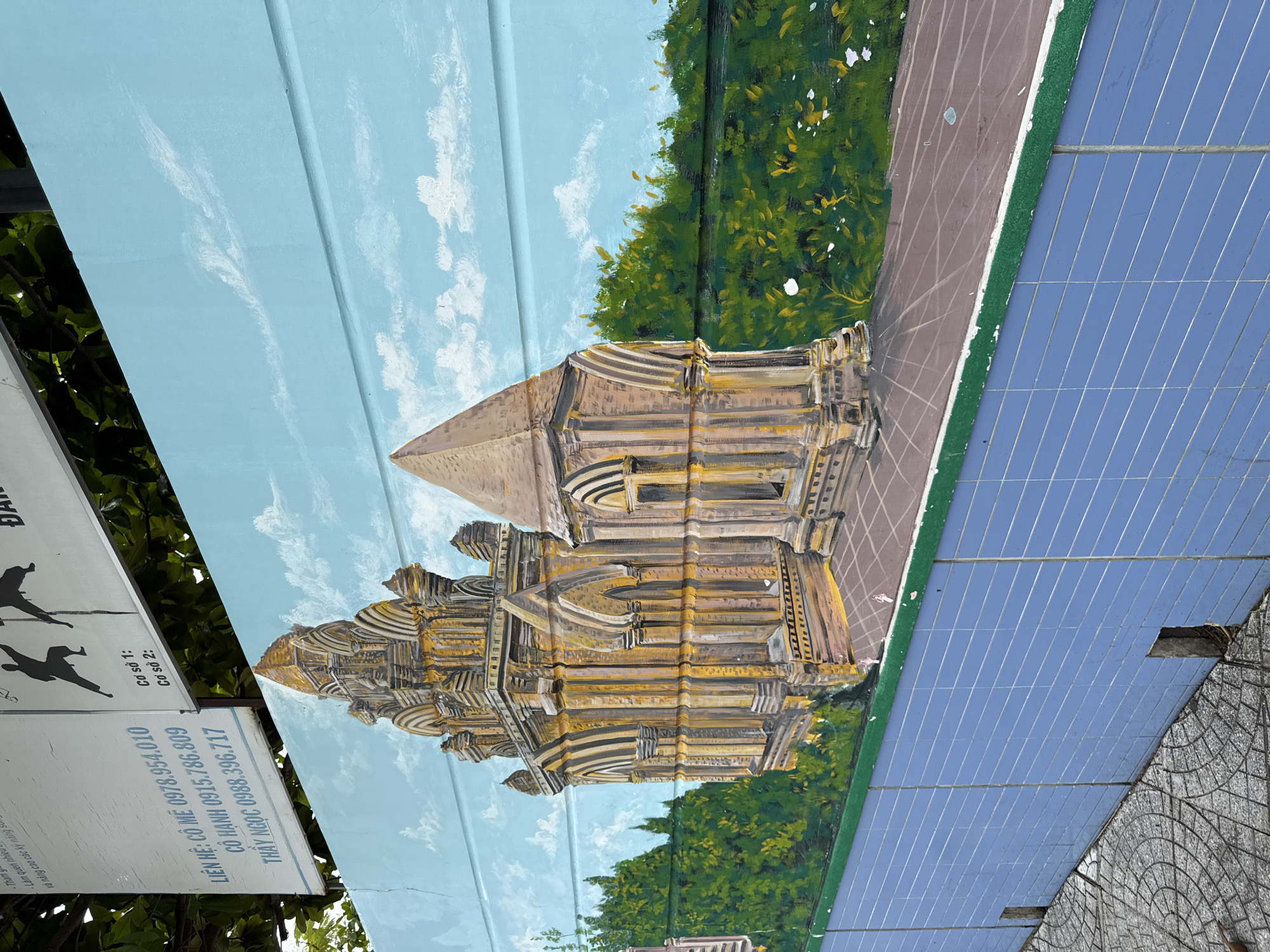 The image depicts a mural painted on a light blue wall with white clouds. The mural features an ornate, ancient-looking stone building with intricate architectural details, rendered in shades of brown and gold. To the left of the building, there is a dense green forest with hints of yellow and white flowers. In the foreground, a grey tiled walkway with circular patterns leads towards the mural. The lower part of the wall is covered in light blue tiles, with two rectangular openings visible at the bottom. Above the mural, a white sign with Vietnamese text and silhouettes of people engaged in martial arts is partially visible. Based on the context of the sign, which mentions "Nha Trang" and a martial arts school, this scene is likely located in Nha Trang, Vietnam, possibly near a martial arts training facility or a public space that features such artwork.