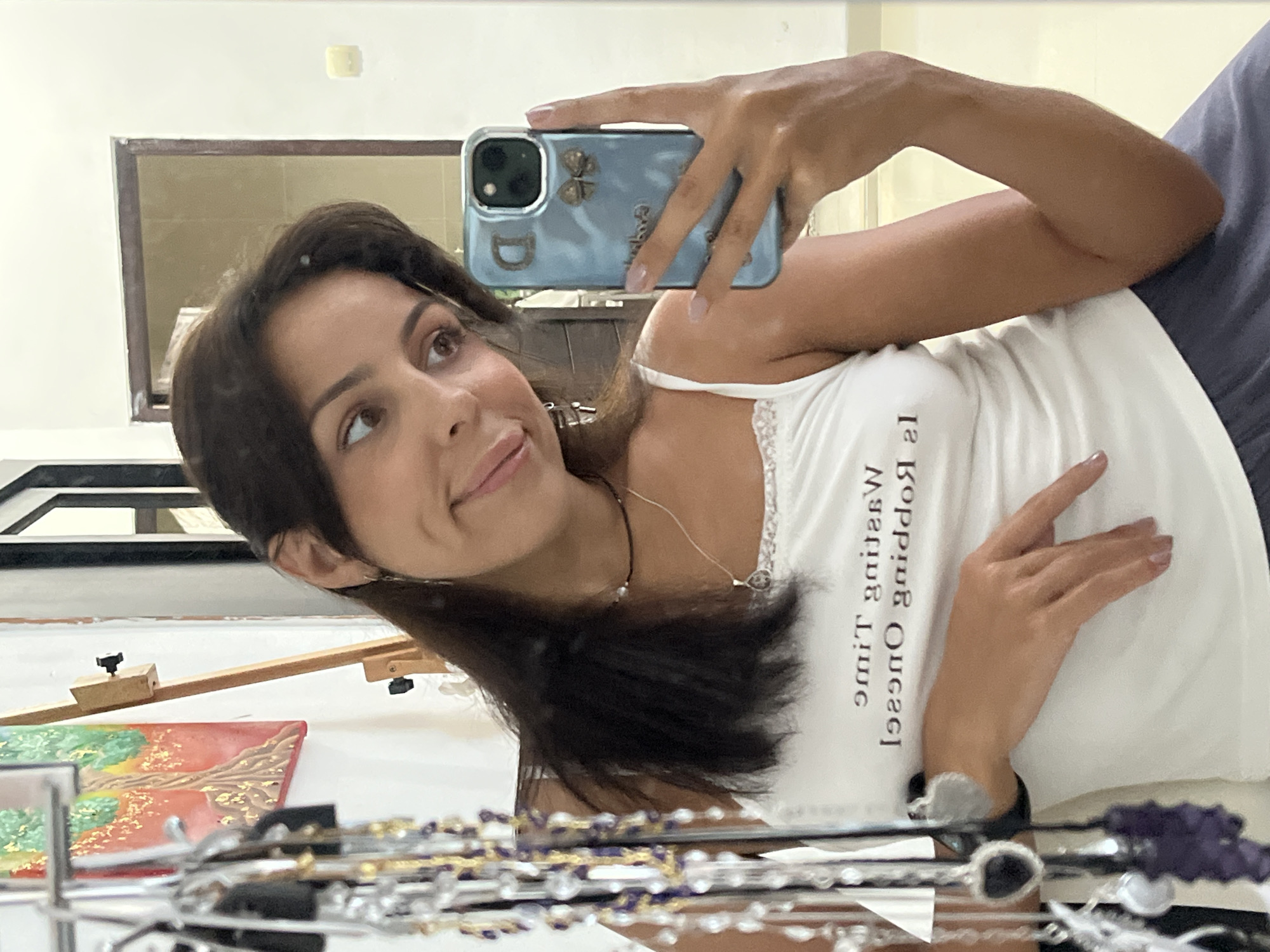 A woman is taking a selfie in a mirror. She is wearing a white tank top with text printed on it and appears to be indoors. The location is Unknown, Indonesia. The reflection in the mirror shows an easel with a painting, and jewelry on display. She is holding a blue smartphone with a decorative case.