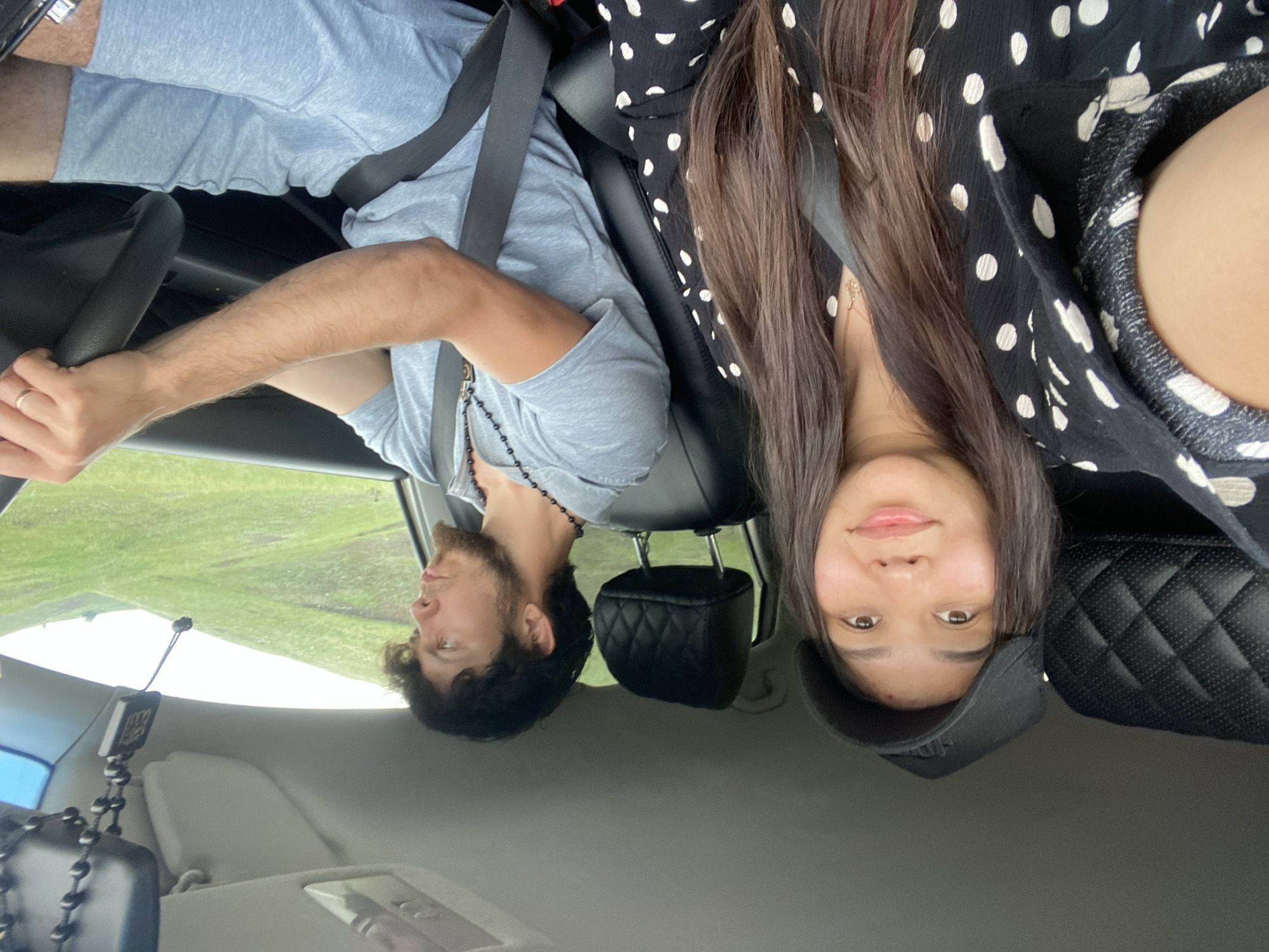 The image shows two people inside a car, likely traveling. A woman with a black baseball cap is in the passenger seat, looking at the camera. She has long dark hair and is wearing a black and white polka dot top. A man is driving the car. He has dark, curly hair and a beard, and is wearing a grey t-shirt and grey shorts. He has a necklace with a cross pendant. They are both wearing seatbelts. Outside the car, green, rolling hills are visible, suggesting a rural or scenic location. There is a beaded religious ornament hanging from the rearview mirror. The interior of the car appears to be a standard passenger vehicle. The scene does not provide any specific clues about the location within the city of Кугарчи, Russia.