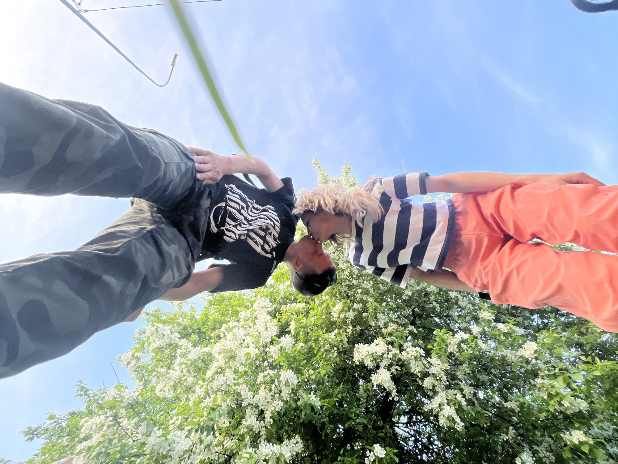 Two people are kissing in Chaikovsky, Russia. One person is wearing camouflage pants and a black shirt with a white design, and the other person is wearing orange pants and a blue and white striped shirt. They are positioned in front of a blossoming tree, with a blue sky visible in the background.