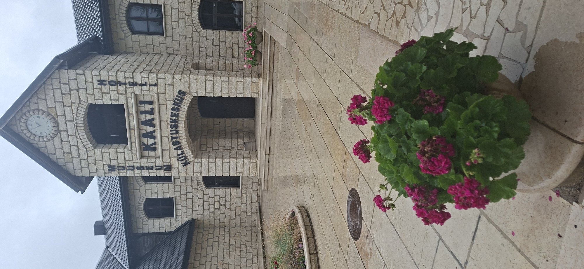 The image shows the Kaali Museum and Hotel in Kaali, Estonia. The building is constructed of light-colored stone bricks and features an arched entryway. Above the entrance, the words "KAALI" are prominently displayed in large, dark lettering. To the left of "KAALI", the word "MUSEUM" is vertically arranged, and to the right, "HOTEL" is also vertically arranged. A circular clock is visible at the peak of the building's facade. In front of the building, a paved courtyard is wet, suggesting recent rain. Potted pink flowers are placed on either side of the building's entrance, and a planter filled with vibrant pink geraniums sits in the foreground of the image. A round metal manhole cover is visible on the wet pavement. The sky is overcast and appears to be raining lightly.