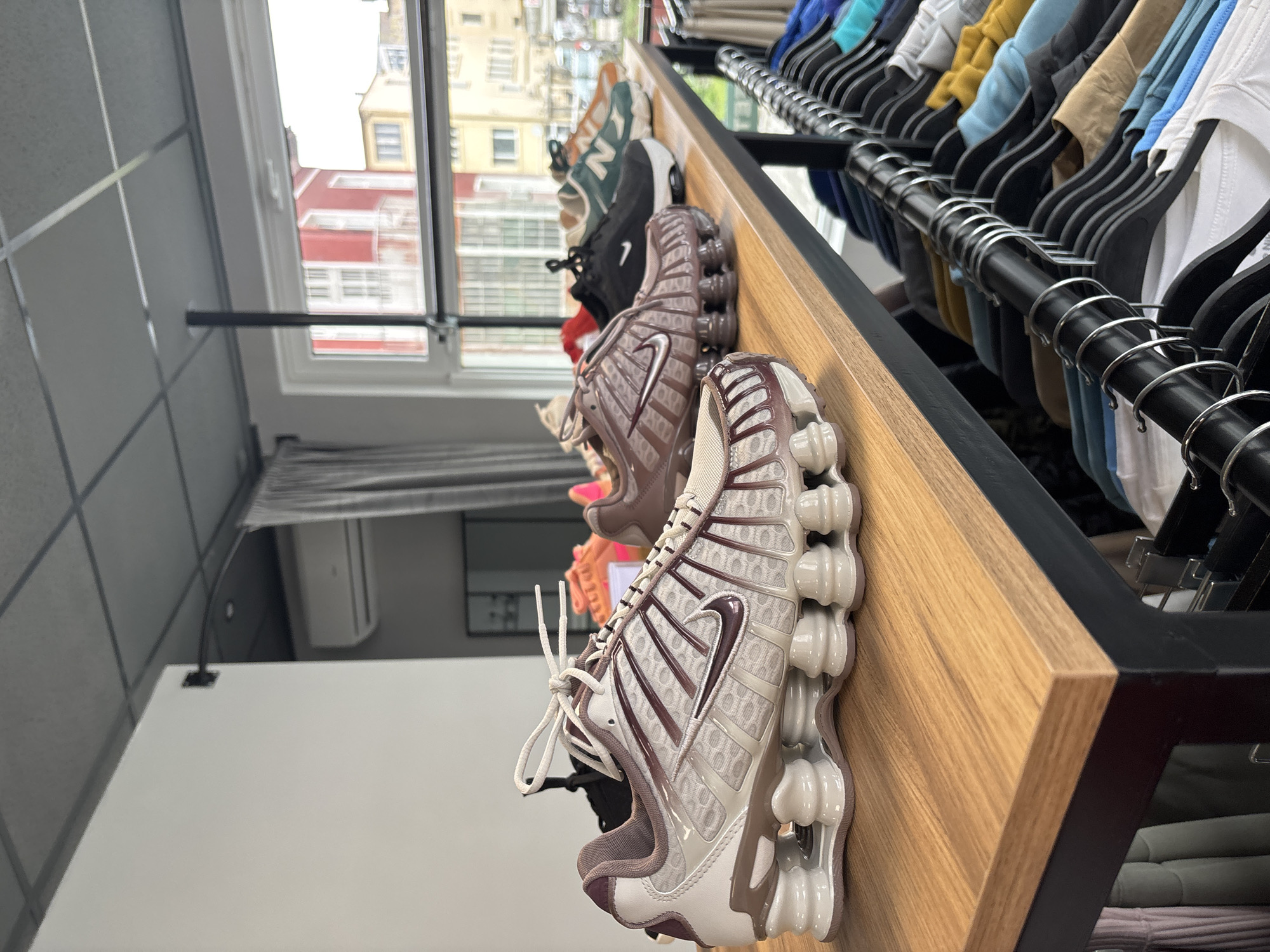 The image shows a selection of athletic shoes displayed on a wooden shelf in what appears to be a retail store. The shoes are in a variety of colors and styles. Behind the shoes, a rack of clothing is visible. Outside the window, a building can be seen. This image was taken in the city of Gabrovo, Bulgaria.
