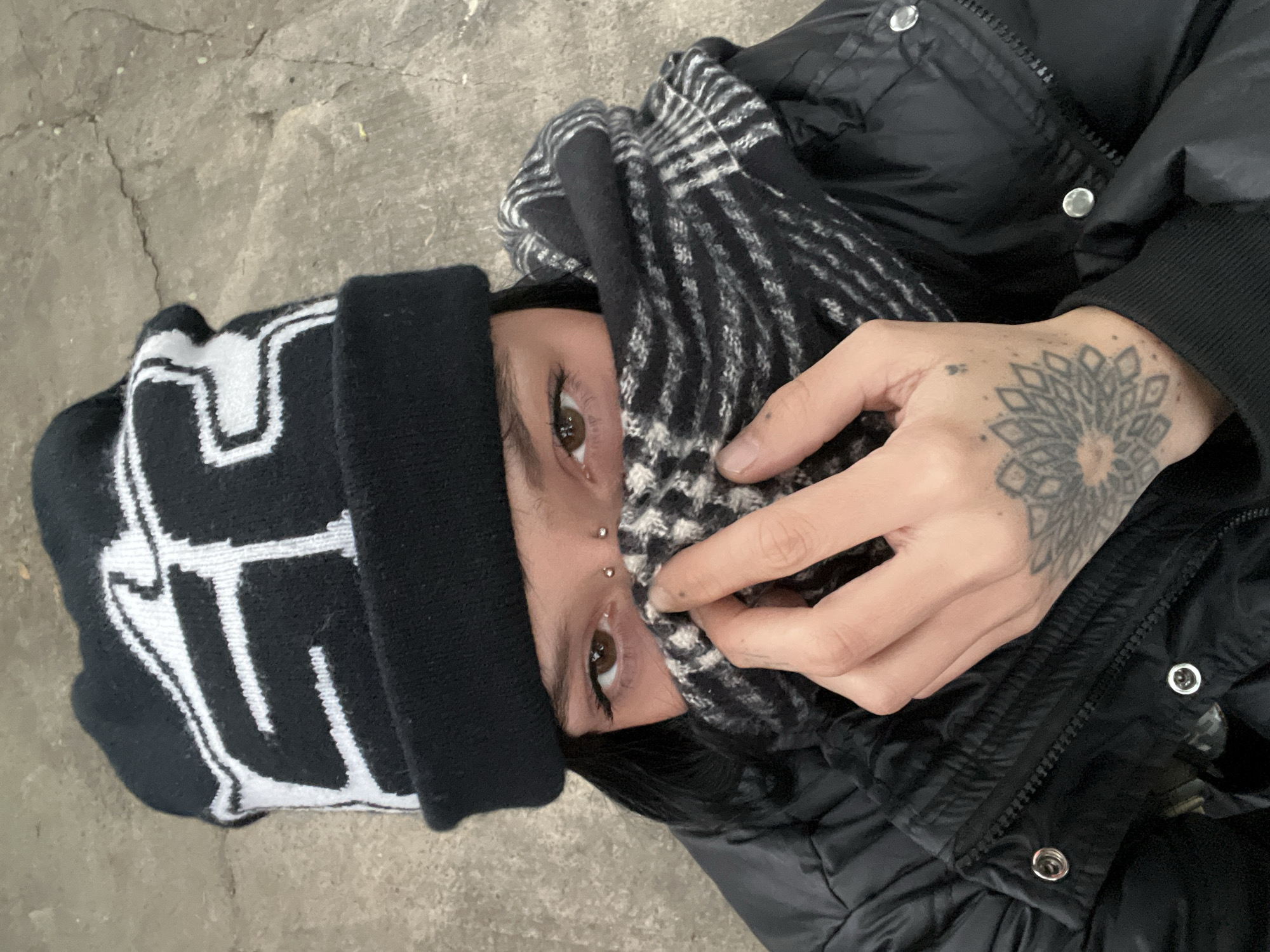 A person wearing a black and white beanie, a black and white scarf, and a black puffer jacket is seen from a high angle perspective. The person has piercings on their face and a tattoo on their hand. The background appears to be concrete. The location cannot be determined from the information provided.