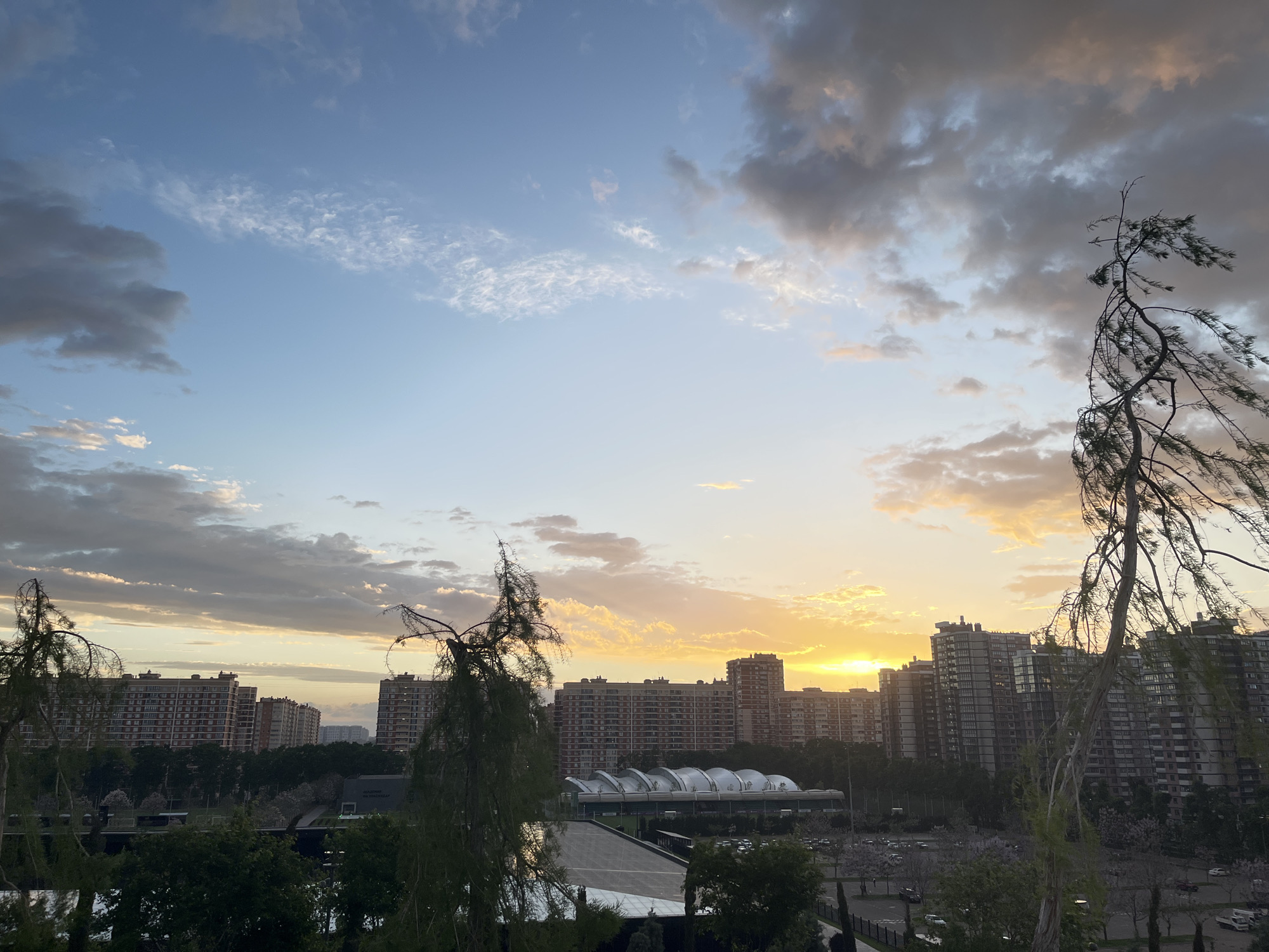 The image shows a cityscape at sunset in Krasnodar, Russia. Multiple high-rise apartment buildings dominate the background, silhouetted against the setting sun. In front of the buildings is a park with trees and a large, arched structure, possibly a sports facility. A few vehicles are visible in a parking area within the park. Two leafless trees frame the view on either side, with branches reaching toward the sky. The sky is a mix of blue and orange, with clouds scattered across the upper portion of the frame.