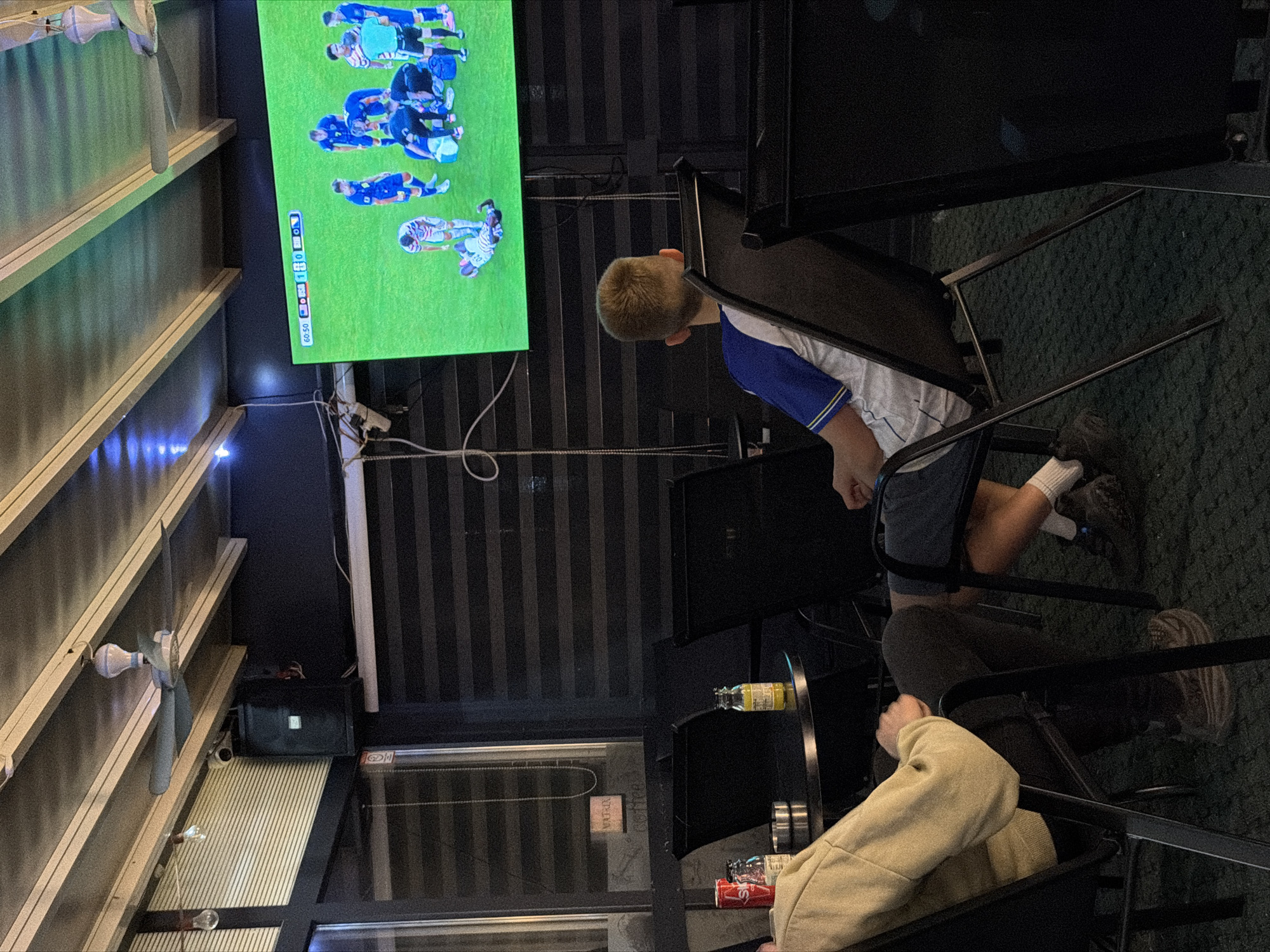The image depicts an indoor, covered seating area, possibly an outdoor cafe or sports bar, in Nević Polje, Bosnia and Herzegovina. A large, flat-screen television mounted on the ceiling displays a live soccer match, with a scoreboard indicating 60:50 minutes played and a score of 1-0. Several individuals are seated, facing the television. In the foreground, a young boy wearing a blue and white soccer jersey is seated in a black chair. To his left, another person is seated, wearing a light-colored hoodie and dark pants. A small round table is positioned between them, holding a red soda can and a small glass bottle. The environment appears to be dimly lit, with striped blinds visible on the walls and ceiling fans overhead. The floor is covered in green carpeting. The overall scene suggests a casual gathering to watch a sporting event.