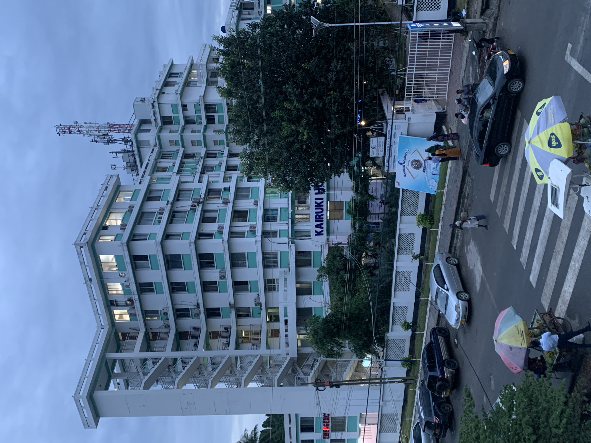 The image depicts a street scene outside the Kairuki Hospital in Dar es-Salaam, Tanzania. The hospital is a multi-story building with a prominent antenna structure on its roof. Several cars are parked along the street. A pedestrian crossing is marked on the road. People are visible on the sidewalk near the hospital entrance and crossing the street. A street vendor with a cart and umbrella is also present. The sky is overcast.