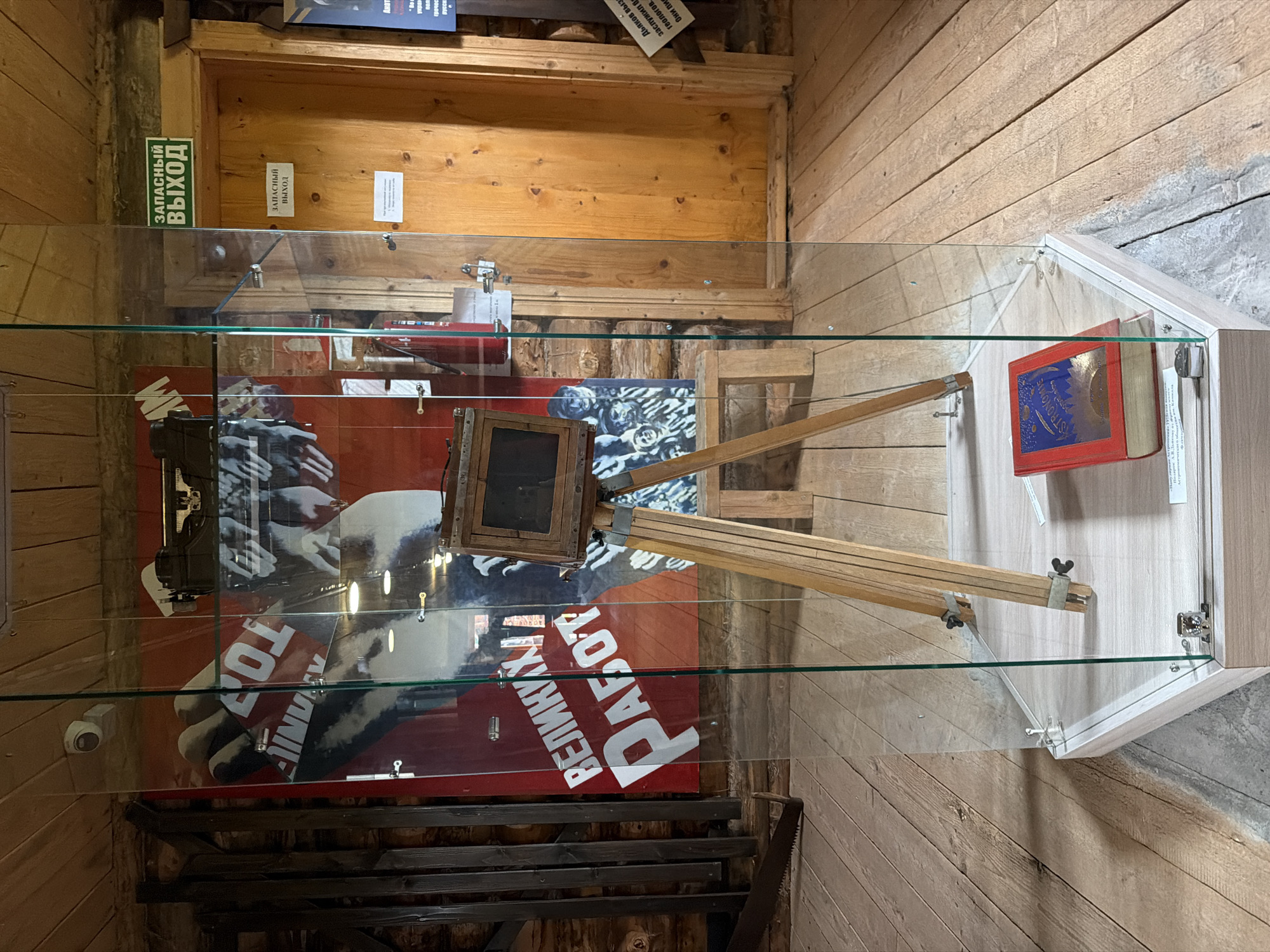 The image shows an indoor exhibit, likely a museum or historical display, featuring a vintage camera on a tripod encased in a glass display case. The camera is a wooden bellows camera with a lens. A red book with gold lettering and a blue insert is placed on the base of the display case. Behind the display case, a large red poster with white lettering and images of hands and faces is visible. To the right of the poster and display case, there is a wooden door with two signs above it, one green and one white, both reading "ЗАПАСНЫЙ ВЫХОД" (Emergency Exit). The surrounding environment consists of wooden walls, a wooden ceiling with visible beams, and a wooden floor. The overall impression is of a rustic, potentially historical setting. The location is identified as Ust'-Pyzas, Russia.