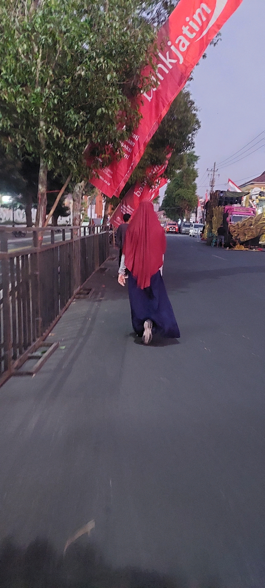 A person wearing a dark blue skirt and a red hijab walks away from the camera on a paved road. A red banner with white text, reading "bankjatim," is visible hanging from trees on the left side of the image. Trees and a fence line the left side of the road. In the background, there are several vehicles, including a pink truck, and buildings. The sky is overcast, suggesting dusk or dawn. The scene appears to be in an urban or semi-urban area in Lumajang, Indonesia.
