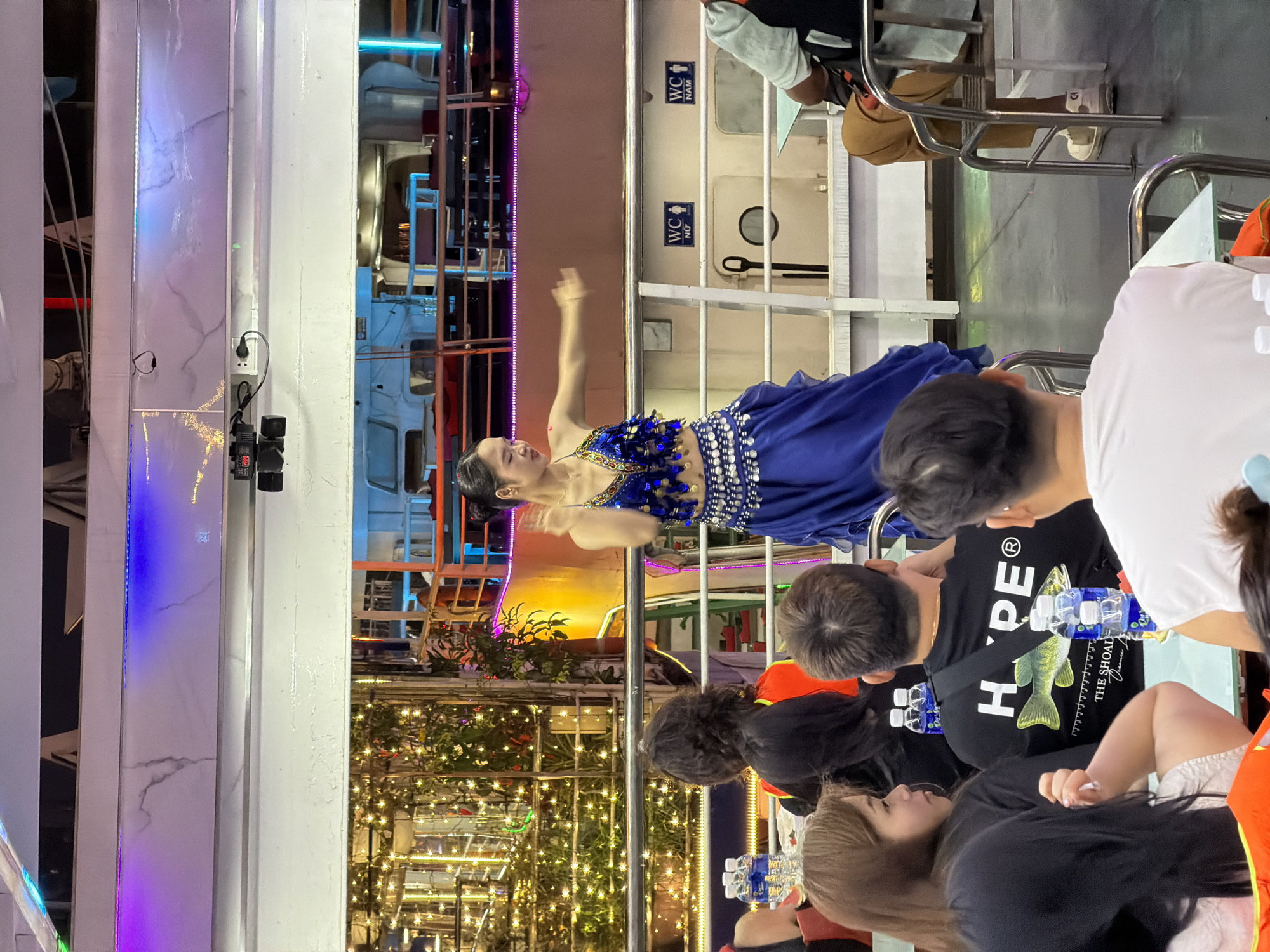 This image captures a vibrant evening scene aboard what appears to be a cruise ship or large tour boat in Đà Nẵng, Vietnam. The central focus is a woman performing a dance, dressed in a striking blue sequined costume with a decorated midriff, suggesting a belly dance or similar cultural performance. Her arms are raised as she dances on a raised platform or walkway.
Several spectators are visible in the foreground, seated at tables and watching the performance. They are predominantly seen from behind or partially, with bottles of water on their tables. One person's black t-shirt clearly displays "HYPE®" and a graphic of a fish with "THE SHOAL" and "DANANG" printed beneath it, strongly confirming the location as Đà Nẵng.
The setting is illuminated by various artificial lights, including strip lights along the walls, fairy lights decorating foliage, and spotlights on the performer, indicating it is nighttime. The interior features white paneled walls, some with a marble-like pattern, and metallic railings. Visible in the background are clear signs for "WC NAM" (men's restroom) and "WC NỮ" (women's restroom), further contextualizing the public venue. An electrical outlet and cables are also seen on a pillar. The overall atmosphere is lively and geared towards entertainment, suggesting a dinner cruise or cultural show experience.