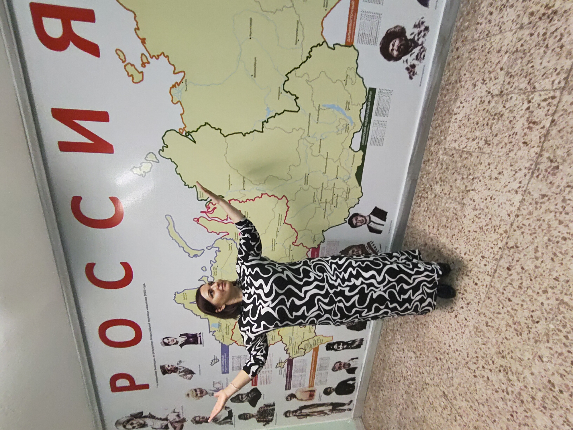 A woman in a black and white patterned dress is standing in front of a map of Russia, presumably in Markovskii, Russia. The map has the word "РОССИЯ" in large red Cyrillic letters on the left side. The woman has her arms outstretched and is smiling. The floor is tiled with a speckled pattern.