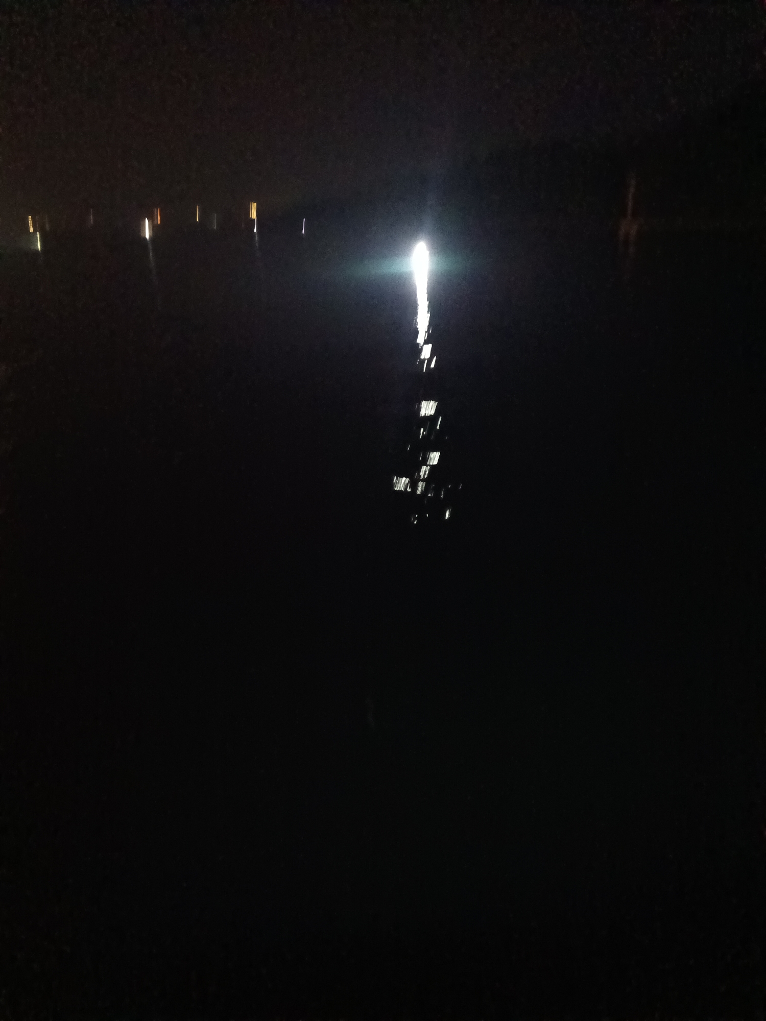 The image depicts a dark, nocturnal scene with reflections on water, likely in Cilegon, Indonesia. A bright light source, possibly a flashlight or camera flash, is prominently featured in the middle of the frame, its reflection extending down into the water. Along the distant horizon, there are several vertical streaks of light, suggesting artificial illumination from buildings or structures. The overall impression is one of a quiet, possibly industrial or waterfront area at night, with the water surface acting as a mirror for the ambient light.