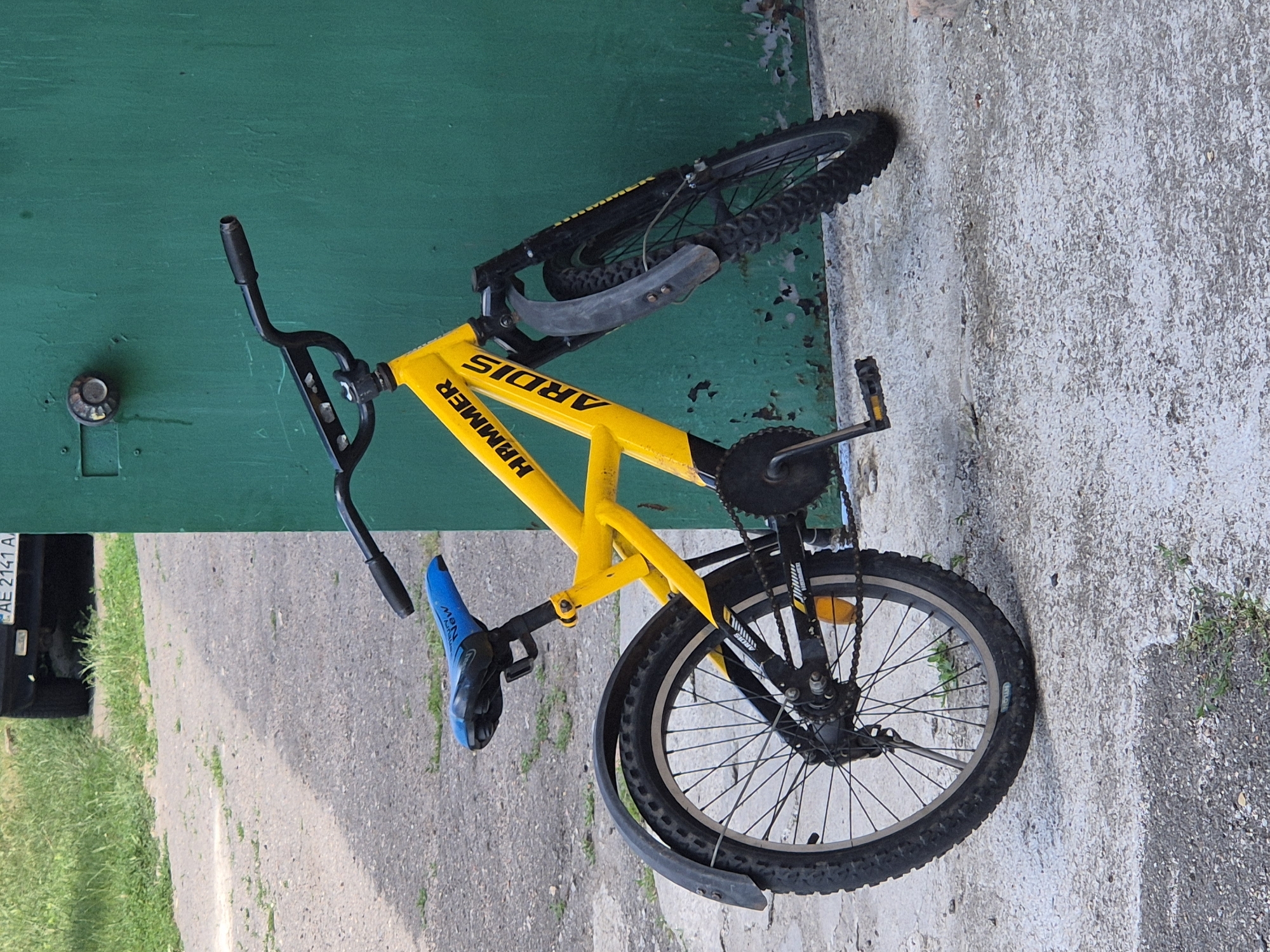 A yellow child's bicycle with black accents is parked against a teal-green metal surface. The bicycle has a blue seat, knobby tires, and fenders on both wheels. Text on the bicycle frame reads "HAMMER" and "ARDIS". The bicycle is on a concrete surface with some gravel and grass. In the background, a portion of a dark-colored vehicle with a license plate that appears to read "AE 2141 A" is visible, along with a patch of green grass. A metal pipe or fixture is attached to the teal-green wall above the bicycle. The scene is outdoors in what appears to be an urban or semi-urban environment, possibly in Sozonivka, Ukraine, given the context provided. There are no people visible in the image.