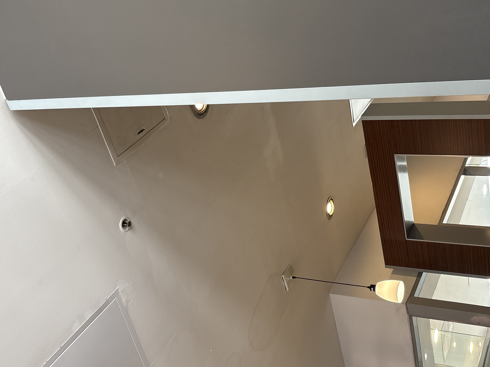 The image shows a view looking upwards towards a ceiling in an indoor environment, possibly a commercial space. In the foreground, a large, flat, gray vertical surface occupies the right side of the frame, obscuring a portion of the scene. The ceiling itself is light-colored and appears to have some water stains or discoloration in various areas.
Several fixtures are visible on the ceiling. A white rectangular panel, likely an access panel or part of the ceiling structure, is present in the upper left. To the right of this panel, a round, white fire sprinkler head is mounted on the ceiling. Further to the right, a smaller, square access panel with a small handle is visible.
In the mid-ground, a pendant light fixture with a white, conical shade hangs from the ceiling by a black cord. Below and to the right of this, a recessed circular light fixture with a warm yellow glow is visible, partially obscured by the gray vertical element.
In the lower portion of the image, a section of what appears to be a wooden frame with a metallic trim is visible. This frame forms an opening, through which a glimpse of a well-lit interior space can be seen, possibly displaying merchandise or an exhibit. Reflections are visible in the glass or reflective surfaces within this opening.
The lighting in the scene is a mix of ambient light and the artificial lights from the fixtures. The overall impression is that of an interior space with standard ceiling finishes and lighting. There are no people or animals visible in the image. The scene does not contain any elements that would specifically indicate a location in Saint Petersburg, Russia.