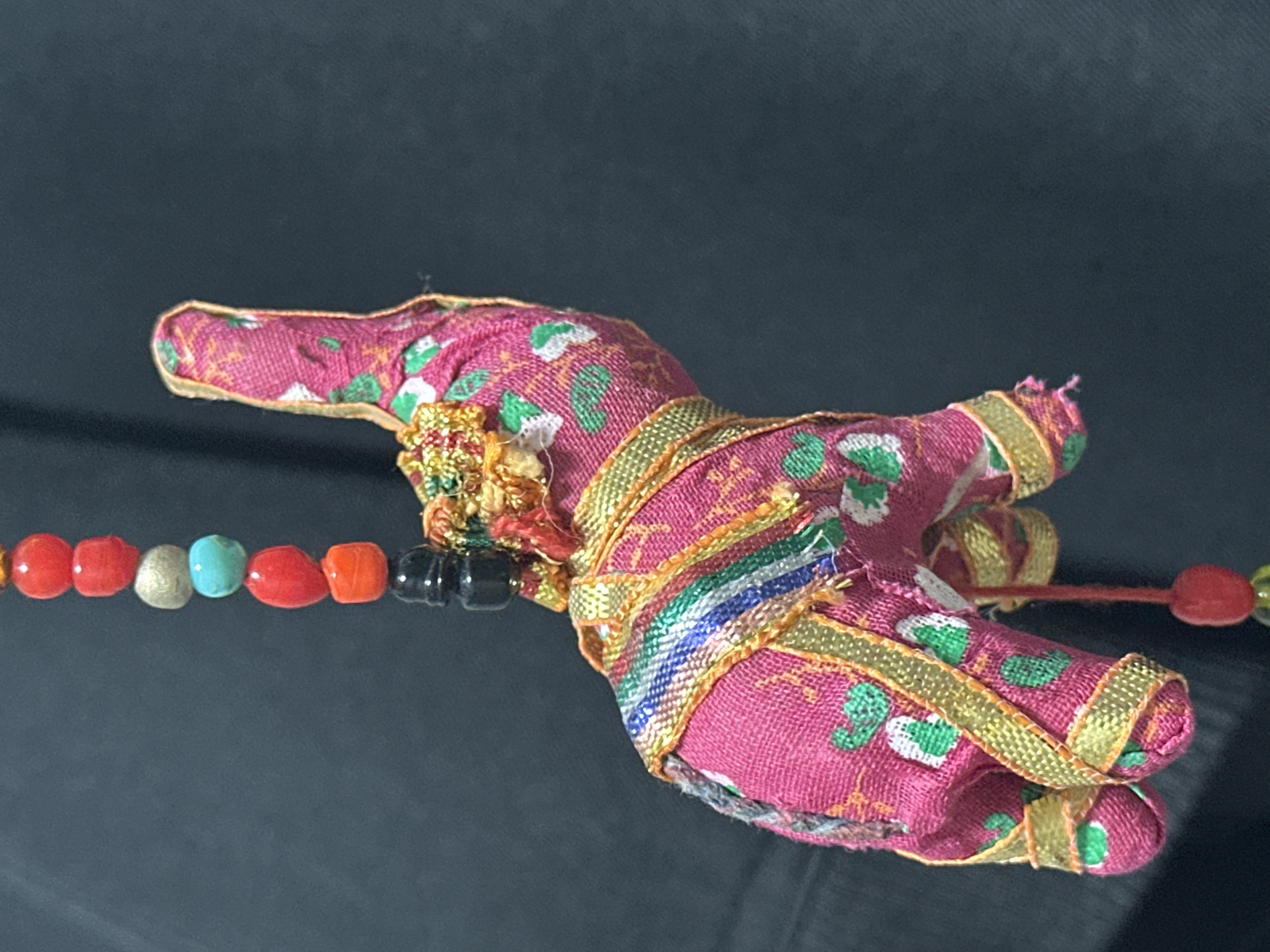 A close-up, eye-level shot shows a brightly colored, fabric-covered elephant ornament hanging against a dark, textured background. The elephant is pink with green and white floral patterns and adorned with gold trim. A strand of colorful beads, including red, silver, turquoise, and black, hangs above the elephant, connected to it by a black bead. The lighting is soft, creating a slight shadow and highlighting the textures of the fabric and beads. The background appears to be a dark fabric, possibly clothing, suggesting the ornament is being held or displayed. There is no indication of the city of Nevinnomyssk, Russia, in the image.