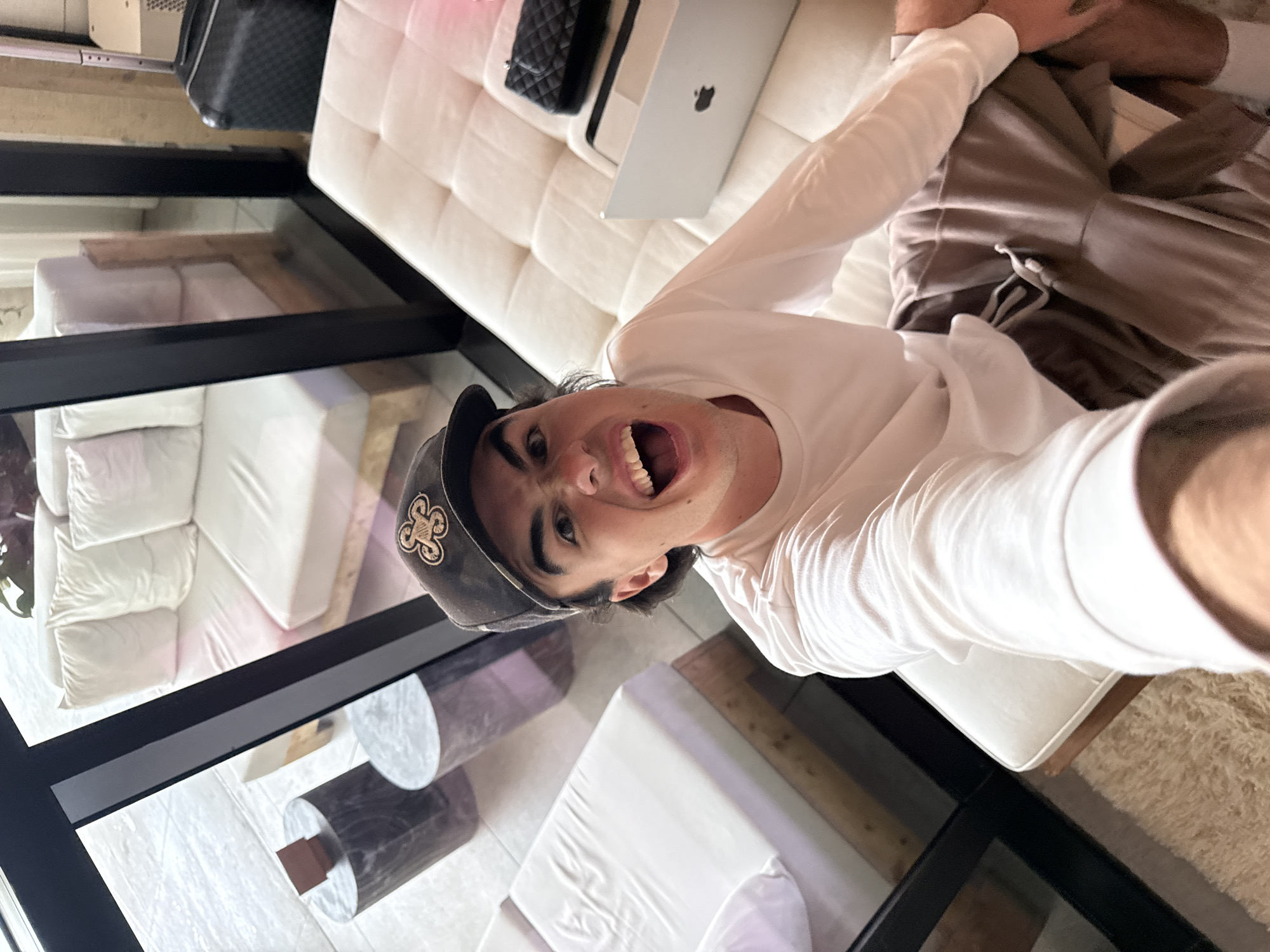 A young man takes a low-angle selfie in a modern, brightly lit interior, likely in Miami Beach, United States of America. He looks up at the camera with a wide-open mouth, conveying excitement, laughter, or a playful shout. He is wearing a white long-sleeve shirt, brown sweatpants, and white socks, and a dark baseball cap with a stylized gold/tan emblem on the front.
The man is positioned on a white tufted sofa or daybed. Next to him on the sofa are a silver Apple MacBook laptop, identifiable by its logo, and a black quilted handbag. In the background, beyond a large glass partition or window, another room is visible, furnished with more white upholstered pieces, possibly beds or sofas. Two cylindrical side tables, one with a light marble pattern and the other dark, are seen through the glass. A black suitcase is partially visible in the upper background. A fluffy beige rug is visible on the floor in the lower right. The overall setting is contemporary and minimalist, with ample natural light suggesting it is daytime.