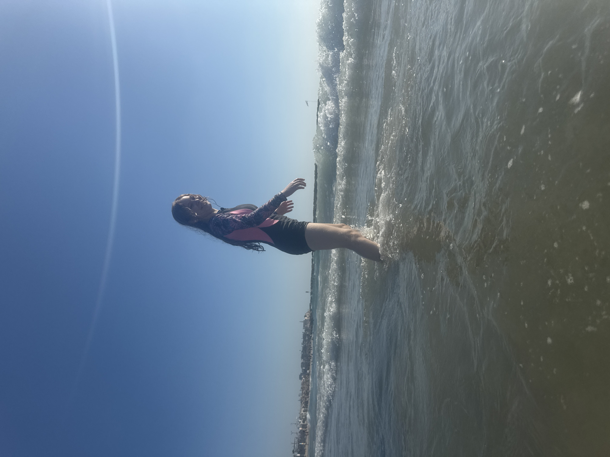 A young woman, wearing a long-sleeved rash guard with pink and black accents, and black shorts, stands in shallow water at the edge of the sea. She is positioned with her back to the camera, looking towards the ocean. Her arms are outstretched to her sides, and her body is angled slightly downwards, suggesting she might be leaning into a wave or balancing herself. The water is clear with gentle waves lapping around her legs, creating some white foam and ripples.
The scene is set on a beach with a clear, bright blue sky dominating the upper portion of the image. A faint, thin white line, possibly a contrail or a light streak, is visible in the sky. In the distance, a coastline with buildings and structures is visible, with the faint outline of the Tel Aviv skyline suggesting the location. The sun appears to be high in the sky, indicating it is daytime. The overall mood is one of leisure and enjoyment of the beach environment. There is no visible text in the image.