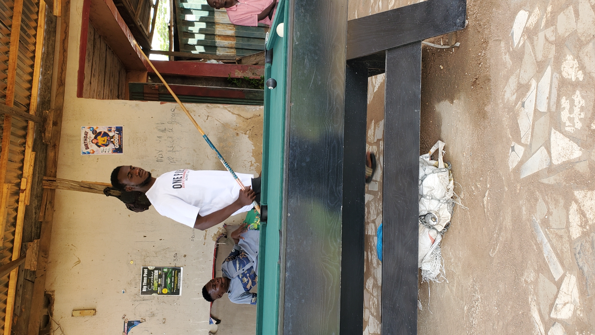 The image depicts an outdoor billiards area, likely in Jalingo, Nigeria, beneath a rudimentary wooden and corrugated metal roof. A pool table with green felt and black wood casing is centrally positioned. Two young Black men are present. One man, wearing a white t-shirt with "ONE PIECE" printed on it, stands to the right, holding a pool cue in mid-action, poised to strike a black billiard ball on the table. He appears to be in his early to mid-twenties with short, dark hair and a beard. To the left, another young Black man, dressed in a patterned grey shirt, reclines on a bench beside the pool table, looking towards the man with the cue. He appears to be in his late teens or early twenties. In the background, a wall exhibits various posters, including one for a "Birthday Party" and another advertising a "Big Time Pool Snooker Competition." The ground is dirt and scattered with stone fragments. The overall impression is of a casual, local recreational space.