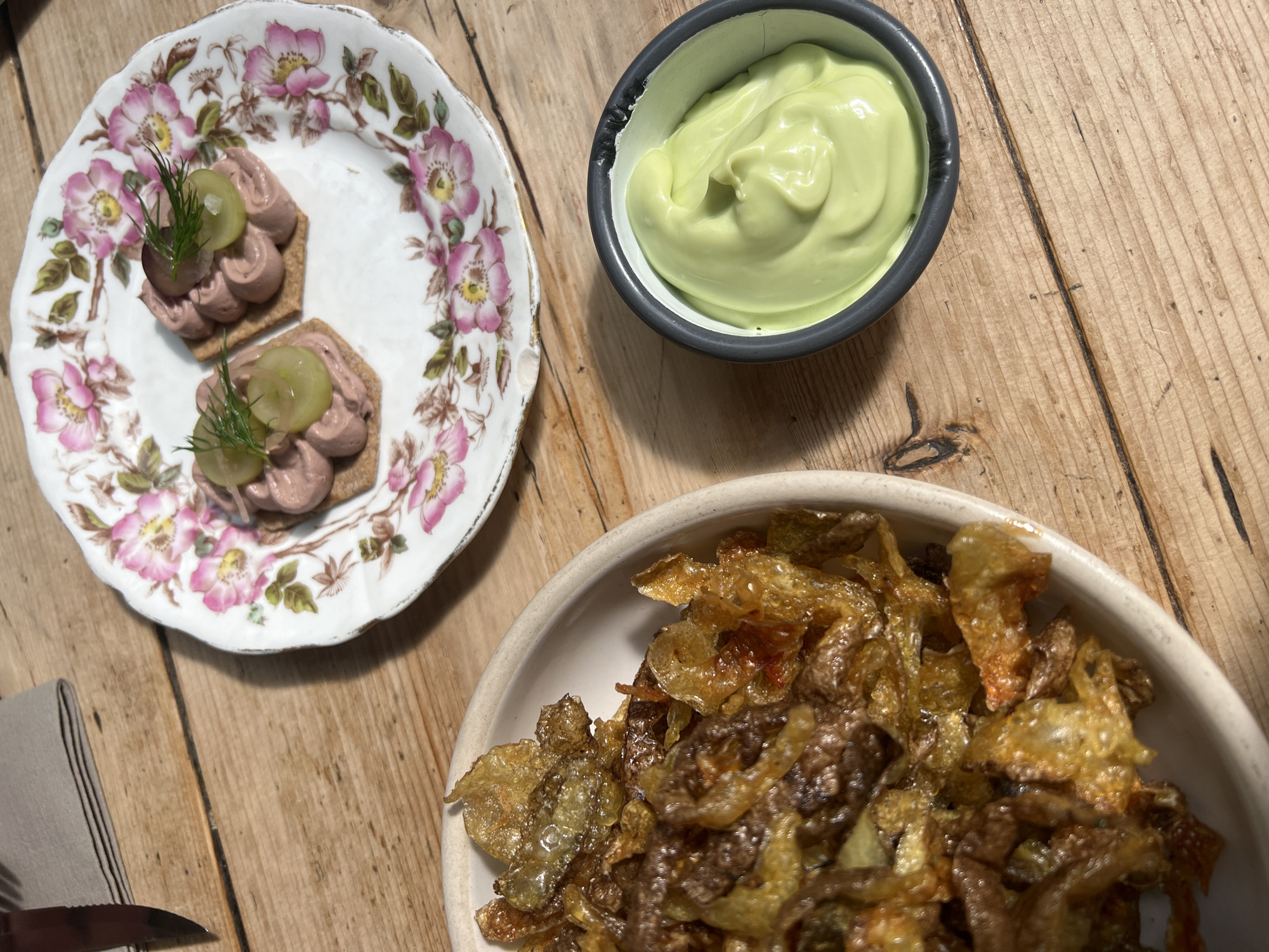 The image depicts a wooden table setting with a plate of hors d'oeuvres, a bowl of what appears to be aioli, and a bowl of fried chips.
The plate contains two small appetizers, each consisting of a square cracker topped with a pâté-like substance, garnished with a slice of grape and a sprig of dill. The plate itself is decorated with a floral pattern.
The aioli is a smooth, light green color and is served in a small, dark-colored bowl.
The bowl of chips contains what appears to be thinly sliced and fried vegetables, possibly potatoes or onions. They have a golden-brown color.
The background is a wooden surface, possibly a table. There is a knife and napkin partially visible in the bottom left corner. The image suggests a food-related setting, such as a restaurant or a home dining arrangement.
There is no explicit information in the image to indicate the location is London, United Kingdom.