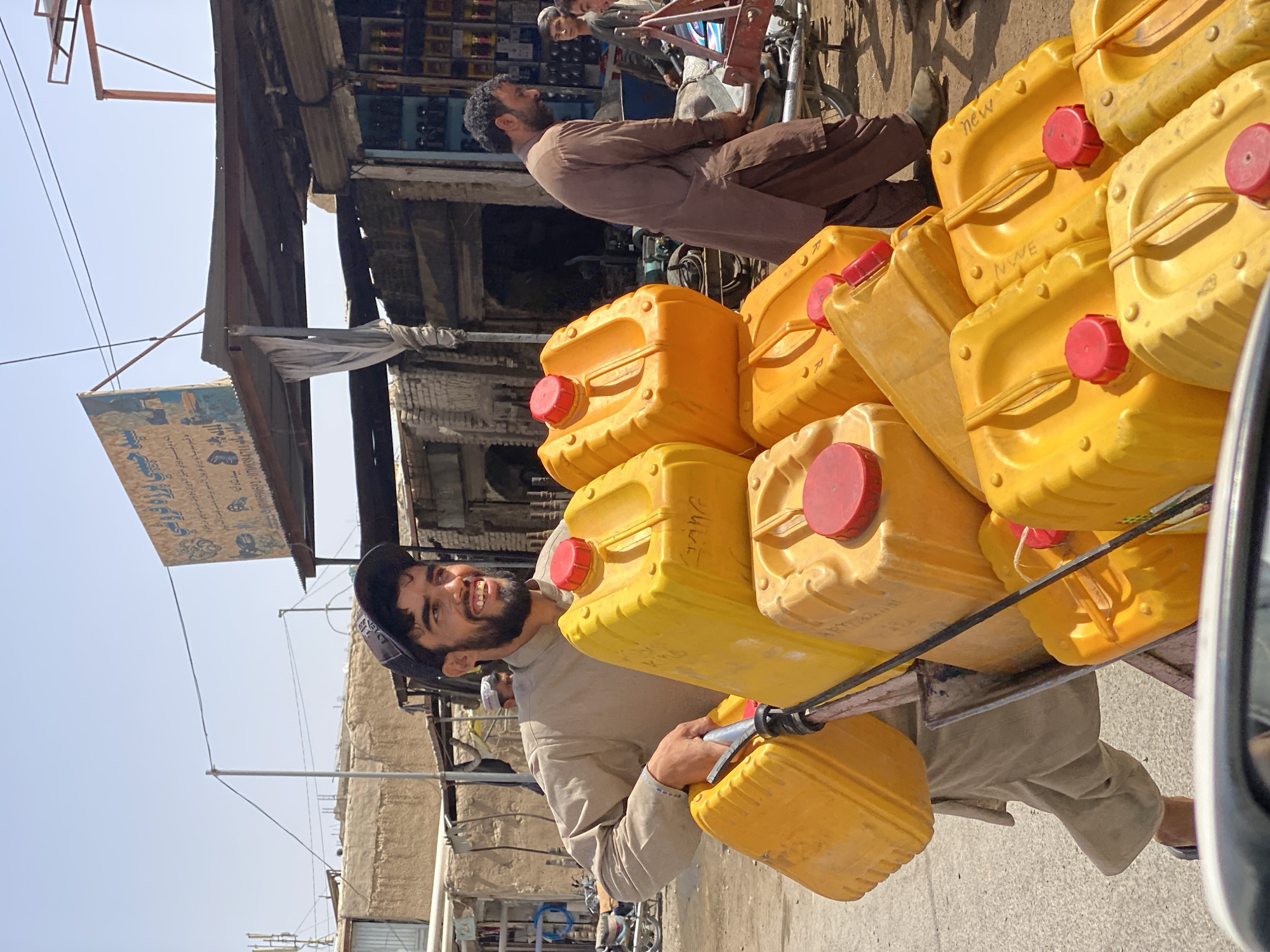 In Kandahar, Afghanistan, a man with a beard and a black baseball cap is pushing a cart loaded with yellow and orange plastic jerrycans. He wears a light brown shalwar kameez. The jerrycans have red caps. Behind him, another man in a dark brown shalwar kameez walks alongside the cart. In the background, market stalls and buildings are visible under a bright, clear sky. A sign with Arabic writing hangs from an awning. The scene appears to be a street in a marketplace.