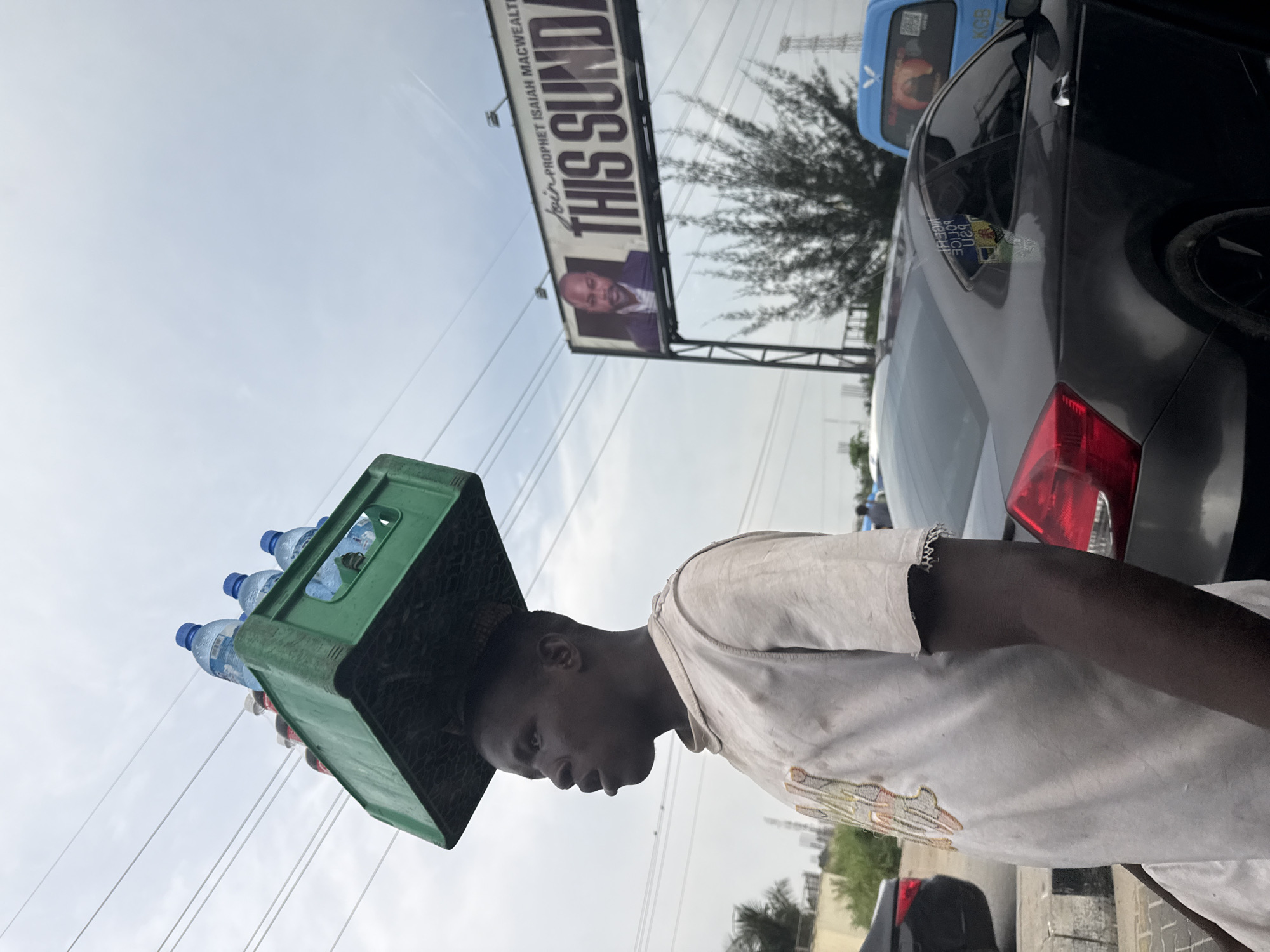 A man carries a green plastic crate of water bottles on his head in Alaguntan, Nigeria. The backdrop features parked vehicles, utility lines, and a billboard advertising Prophet Isaiah Macwealth with the text "THIS SUNDAY." The man wears a light-colored shirt and a bracelet.