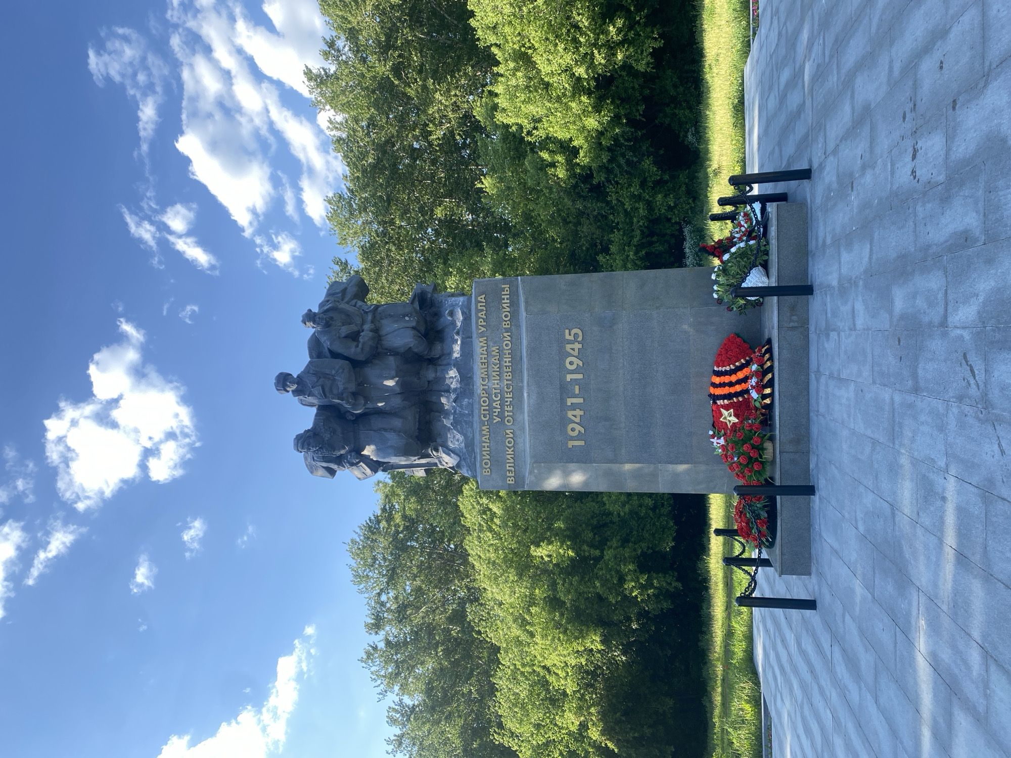 The image depicts a monument in Yekaterinburg, Russia dedicated to the soldiers from the Ural region who participated in the Great Patriotic War (World War II). The monument features a bronze sculpture of soldiers atop a gray stone plinth. The plinth is inscribed with the years "1941-1945" in gold lettering and some text in Russian. Wreaths of flowers, including some with red and orange ribbons, are placed at the base of the monument. The monument is situated on a gray brick or stone surface, bordered by green grass and trees. The sky is blue with scattered white clouds.
