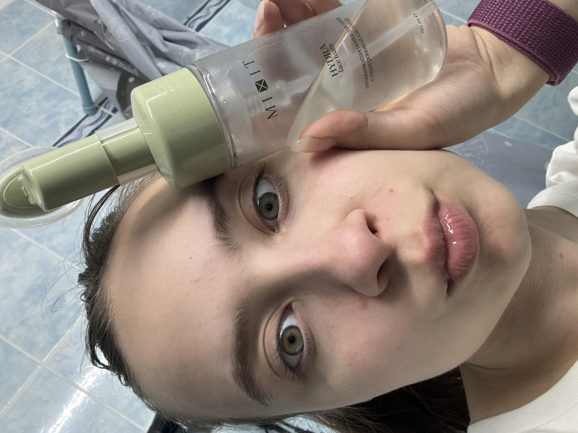 A close-up, slightly rotated photograph shows a young woman holding a bottle of "MIXIT Hydra Face Foam" against her forehead. She is gazing upwards towards the camera with wide, hazel eyes. The bottle is clear with green accents, and the label is partially visible, indicating it contains 150 ml of product. The woman's skin appears clear, and her lips have a glossy finish. A purple hair tie is visible on her wrist. The background consists of blue and white tiles, suggesting a bathroom setting. A small portion of what appears to be a draped cloth is visible in the upper left corner. There is no specific indication of the location being Прикамский, Russia, from the visual evidence.