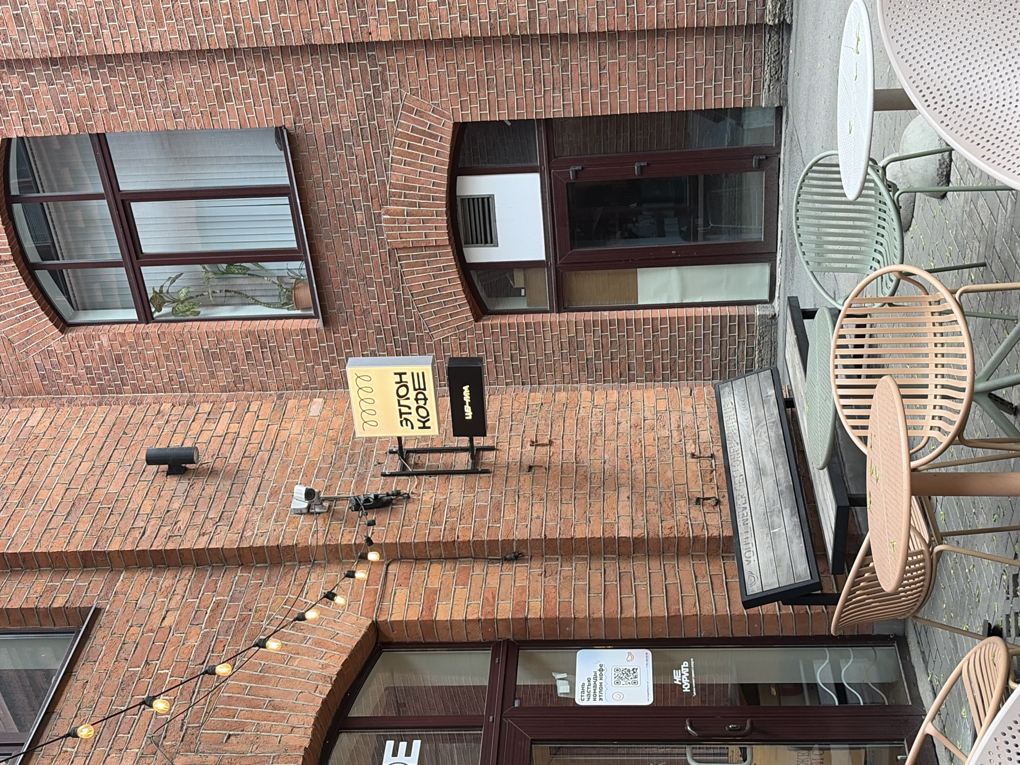 This image depicts the exterior of a cafe in Saint Petersburg, Russia, likely located in a courtyard or alleyway given the brick architecture and limited view. The cafe, named "ЭТЛОН КОФЕ" (Etlon Coffee), is identified by a sign above its entrance, which is a brown door with glass panels. A smaller sign on the door indicates "НЕ КУРЯТЬ" (No Smoking).
The cafe has an outdoor seating area with several tables and chairs. There are light pink, pale green, and white chairs arranged around round tables. One table is covered in leaves. A wooden bench with a metal frame is positioned against the brick wall. Above the bench, the inscription "YOU NEVER GET LONGER THAN YOU NEED" is visible.
The building itself is constructed of red brick, with arched windows and exposed brickwork detailing. Several string lights are hung along the wall near the entrance, adding a decorative touch. A security camera is mounted on the wall. The ground is paved with interlocking bricks. The overall impression is of a cozy, urban cafe setting.