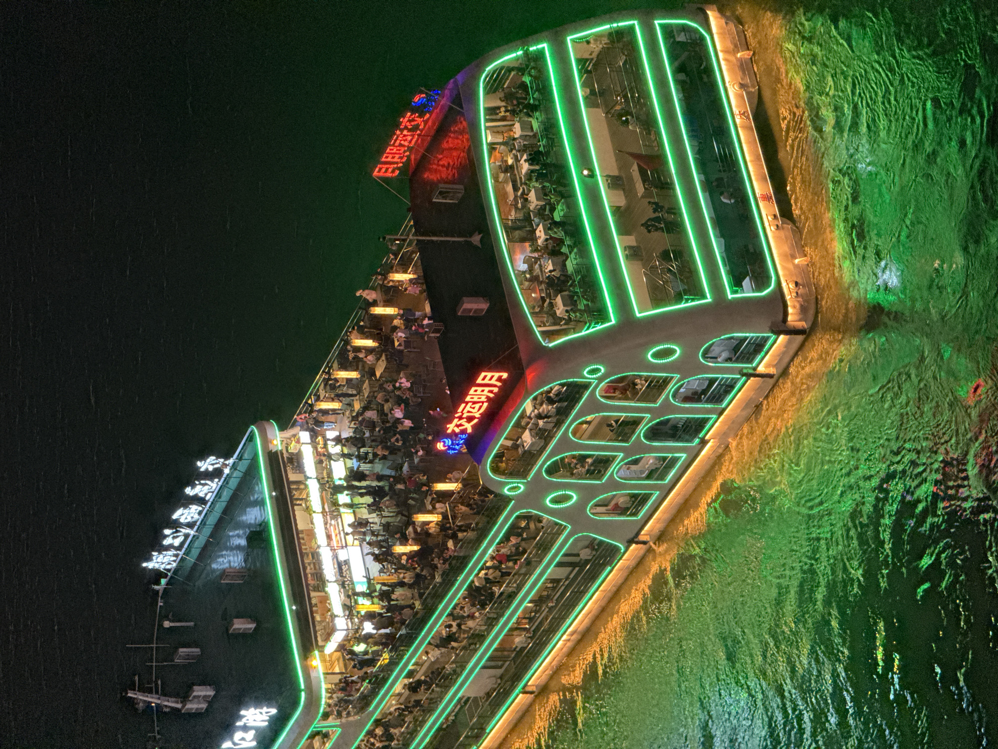 The image captures an illuminated, multi-level boat, seemingly a dinner cruise or leisure vessel, navigating a body of water at night. The boat is crowded with people, mostly seated at tables on multiple decks. Bright green neon lights accentuate the boat's structure, particularly around the edges of the decks and windows. The water reflects the green light, creating a vibrant, almost surreal effect. Chinese characters are visible in neon lighting on the boat's facade. The scene is likely located in the Yuzhong District, China, given the context.