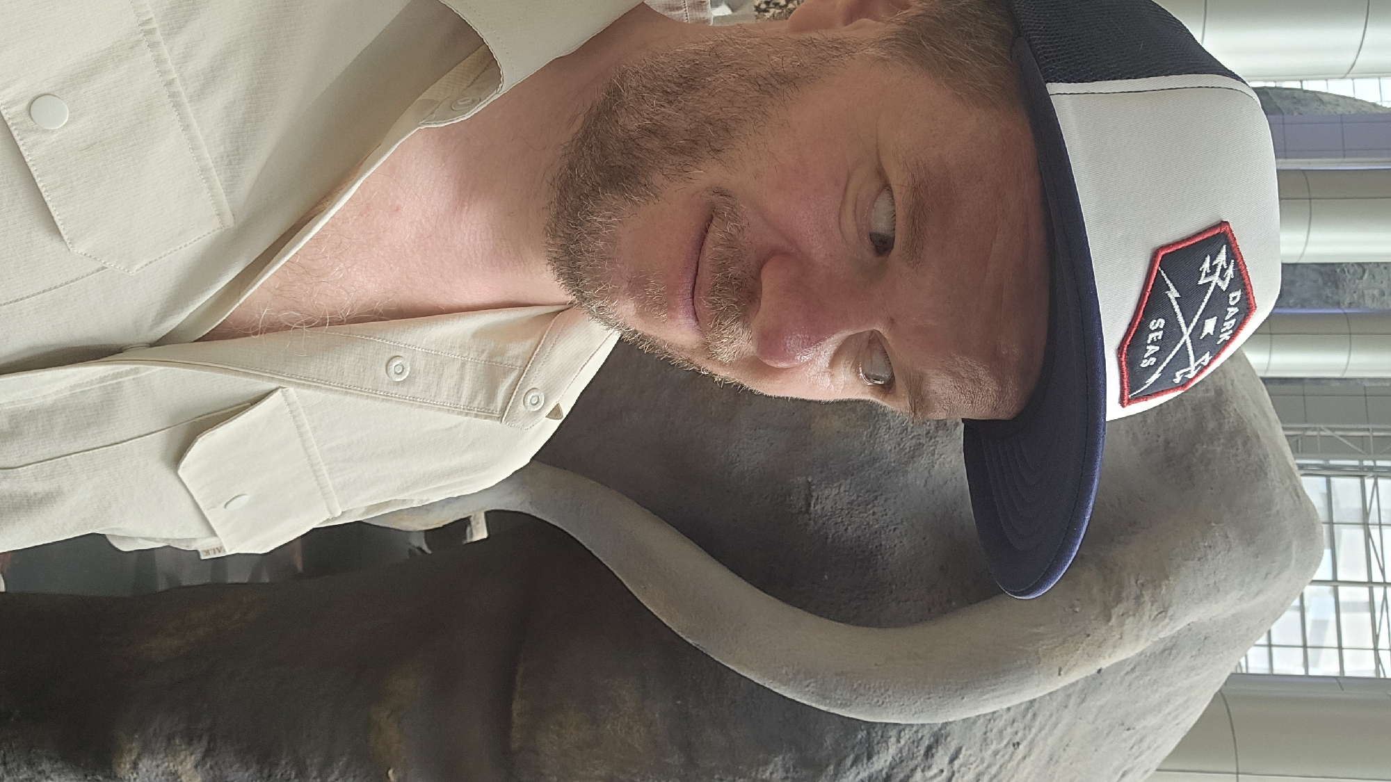 A man wearing a white and navy trucker hat with a patch that reads "DARK SEAS" is seen from the chest up. He has light skin, a light brown beard, and is looking to his right. He is wearing a light beige button-up shirt with two chest pockets. Behind him, to his right, is the textured gray side and tail of a large elephant statue or exhibit. The background consists of tall, cylindrical pillars and a window with a grid pattern. This scene is likely located in the Gongshu District of China, possibly within a museum or exhibition hall.
