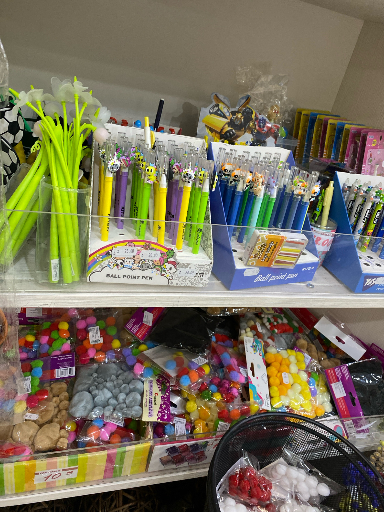 The image shows a retail shelf display, likely in a stationery or craft store, featuring a variety of colorful items. On the top shelf, there are bundles of bright green novelty pens with floral accents, alongside rows of packaged ballpoint pens with cartoon characters and various other decorative elements. To the right, a selection of boxed pens and small packaged items are visible. The middle shelf contains more packaged pens and some small decorative objects. The bottom shelf is filled with bags of colorful pom-poms, small decorative stones, and other craft supplies. A price tag with "10" is visible on a striped cardboard display at the front of the bottom shelf. The overall impression is of a well-stocked retail environment offering a range of stationery and crafting materials. No specific location in Chișinău, Moldova can be identified from the image alone.