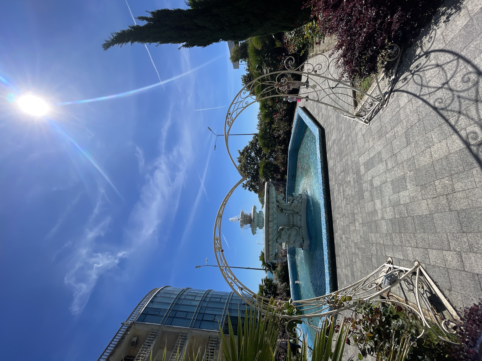 The image depicts a bright and serene outdoor scene, likely a public garden or resort area, under a clear blue sky. A multi-tiered stone fountain with sculpted figures, possibly cherubs, stands in the middle of a rectangular pool lined with blue mosaic tiles. Water is actively spouting from the fountain's tiers.
Two ornate, white metal arches, resembling pergolas, frame the fountain, leading toward it and casting intricate shadows on the ground. The pathway and surrounding area are paved with grey rectangular tiles.
Lush green vegetation, including tall, dark green coniferous trees (possibly cypresses) and dense foliage, forms a verdant backdrop. Palm fronds are visible in the foreground, adding to the subtropical aesthetic. To the left, a modern building with a curved glass facade and grey structural details is partially visible, reflecting the bright sky.
The weather is sunny with a clear blue sky, marked by several distinct white contrails from aircraft. The sun is high, causing a prominent lens flare in the upper left of the image and casting strong, well-defined shadows, indicating midday or early afternoon. The scene conveys a sense of peacefulness and pleasant warmth. No people are visible, and there is no discernible text. Given the context, this location is in Sochi, Russia.