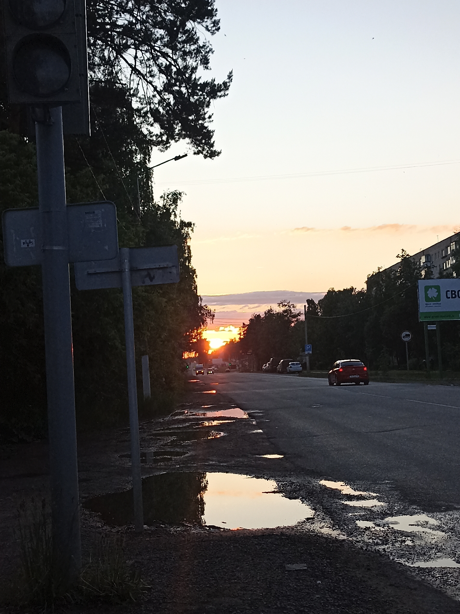 The image captures a roadside scene at sunset in Chaikovsky, Russia. The sun, a bright orange and yellow orb, is setting low on the horizon, partially obscured by distant trees, and casting a warm glow across the sky. The foreground is dominated by the silhouetted back of a traffic light and several sign poles, with one sign displaying faint "ROS" lettering. The left side of the frame features dense, dark green trees and bushes, silhouetted against the bright sky.
Numerous puddles of varying sizes are scattered along the dirt shoulder and on the asphalt road, reflecting the vibrant sunset colors. These puddles indicate recent rain. The road itself appears somewhat worn, with visible potholes.
In the mid-ground, a red hatchback car is driving away from the viewer on the two-lane road, its brake lights possibly illuminated. Other indistinct cars are parked further down the road on the left side. A blue road sign with a white "P" for parking is visible on the right side of the road.
Towards the right, a large billboard with a green background stands prominently. It features a four-leaf clover logo and clearly displays the Russian text "СВО" and "СберСпасибо" (SberSpasibo), indicating an advertisement for Sberbank's loyalty program. Behind the billboard, a multi-story residential building with visible balconies stretches across the upper right part of the frame, also partly silhouetted.
The overall mood is tranquil, with the soft light of dusk highlighting the reflections and silhouettes. The sky above is a gradient of pale blue to warm orange and pink near the horizon.
