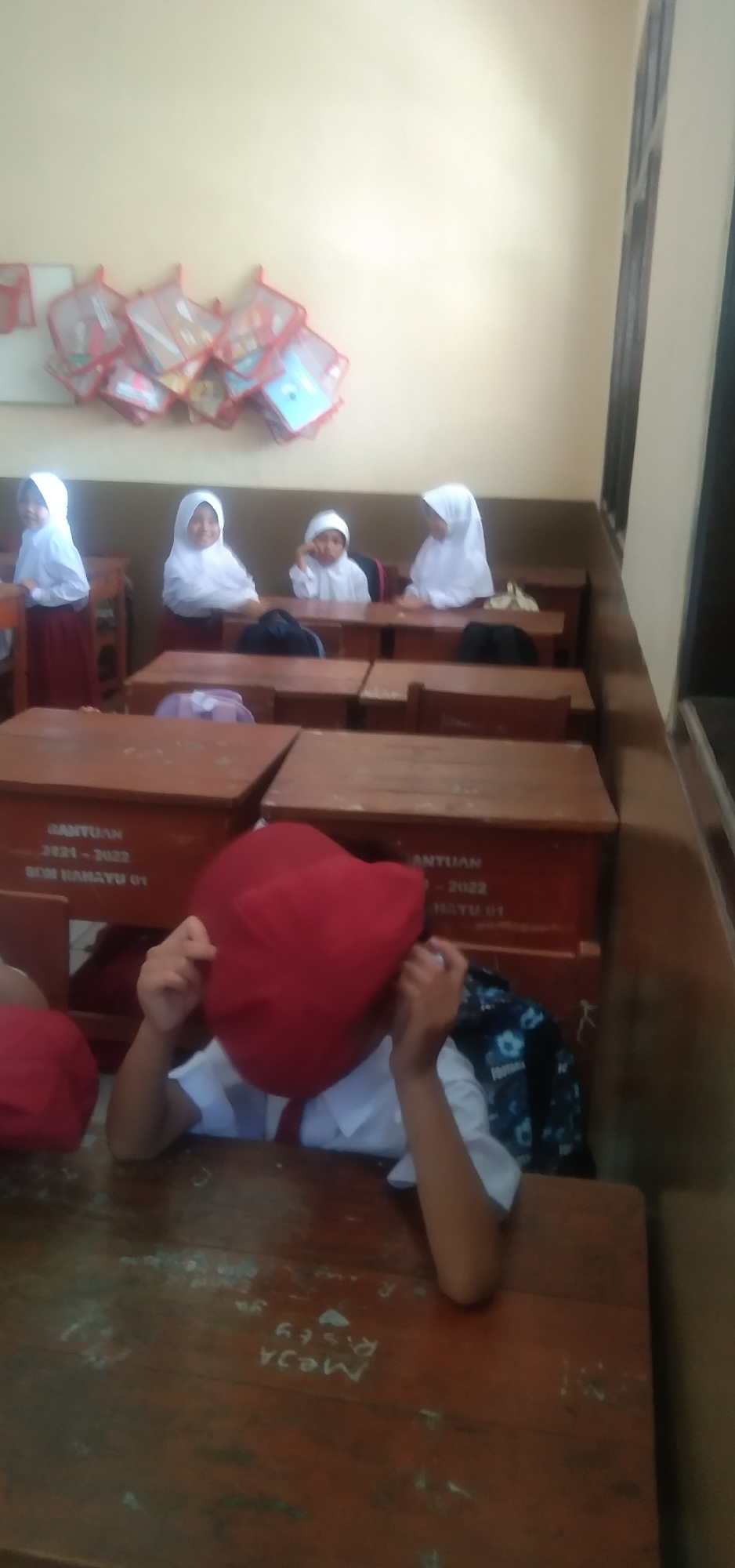 In a classroom in Rahayu, Indonesia, four young girls in white hijabs and white uniforms with red skirts are seated at wooden desks. The girl in the foreground is wearing a red cap that obscures her face and is resting her head on her hands. She is wearing a white shirt and a red tie. Behind her, other students are seated at their desks. On the wall above the students, several mesh bags containing colorful items, possibly books or school supplies, are hung. The classroom appears to be dimly lit, with natural light entering from an unseen window. The desks have markings scratched into the wood. The overall scene depicts a typical classroom setting.