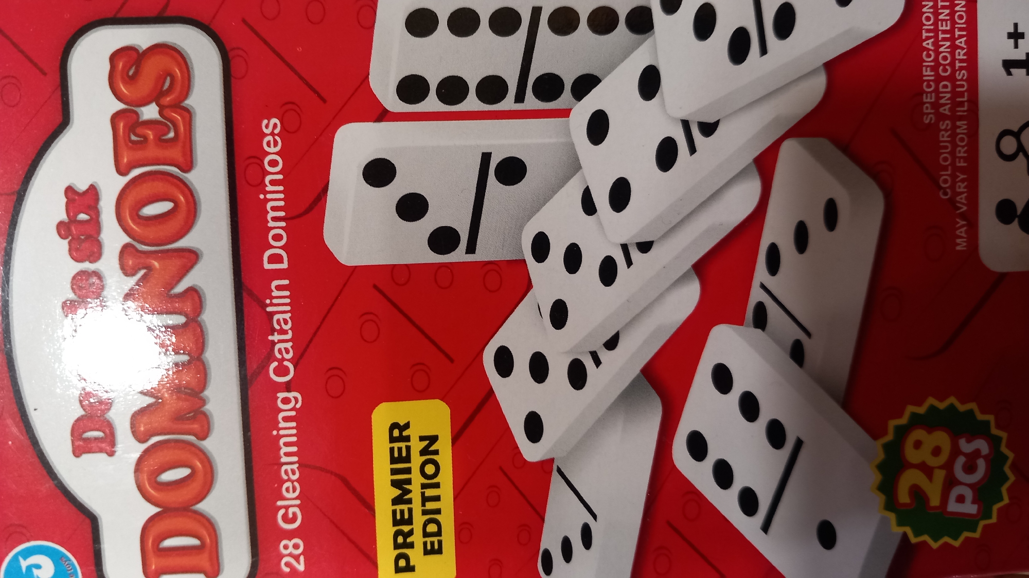 The image is a product shot of a dominoes game box. The box is predominantly red and features the brand name "DOMINOES" in large, orange, 3D lettering at the top. Below the brand name, it reads "28 Gleaming Catalin Dominoes." In the upper left corner of the box, there is a yellow sticker with the text "PREMIER EDITION." The image also shows several white dominoes with black dots, arranged in a fanned-out pattern across the bottom half of the box. The background of the box has a subtle pattern of concentric circles and diagonal lines. In the lower left corner of the box is a circular emblem with the number "28" and the letters "PCS" inside. The bottom right corner has text that reads "SPECIFICATIONS COLOURS AND CONTENT MAY VARY FROM ILLUSTRATION" and an age recommendation icon that shows "1+". The image does not provide any information about the city of Kos, Greece, or any specific location within it.