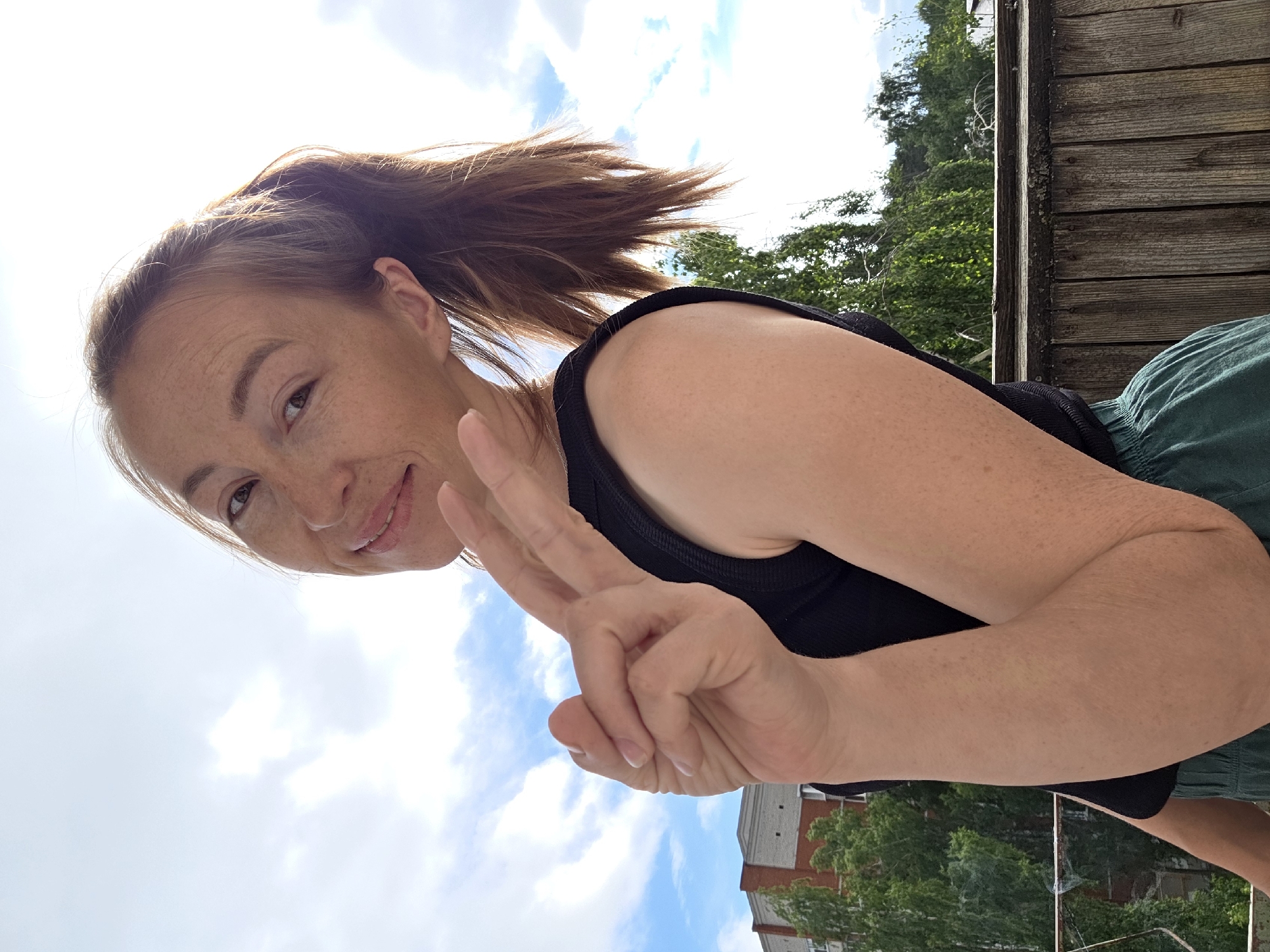 In this medium shot, a woman with light skin and freckles stands outdoors, her body angled to the right. She has her hair pulled back into a ponytail and is wearing a black tank top and green shorts. Her right arm is bent, and she is holding up two fingers in a peace sign. The background shows a cloudy sky with some blue peeking through. To the left, a brick building and green trees are visible. To the right, a wooden fence runs horizontally across the lower portion of the image. The overall scene suggests a casual, outdoor setting, possibly on a balcony or rooftop in the city of Chaikovsky, Russia.