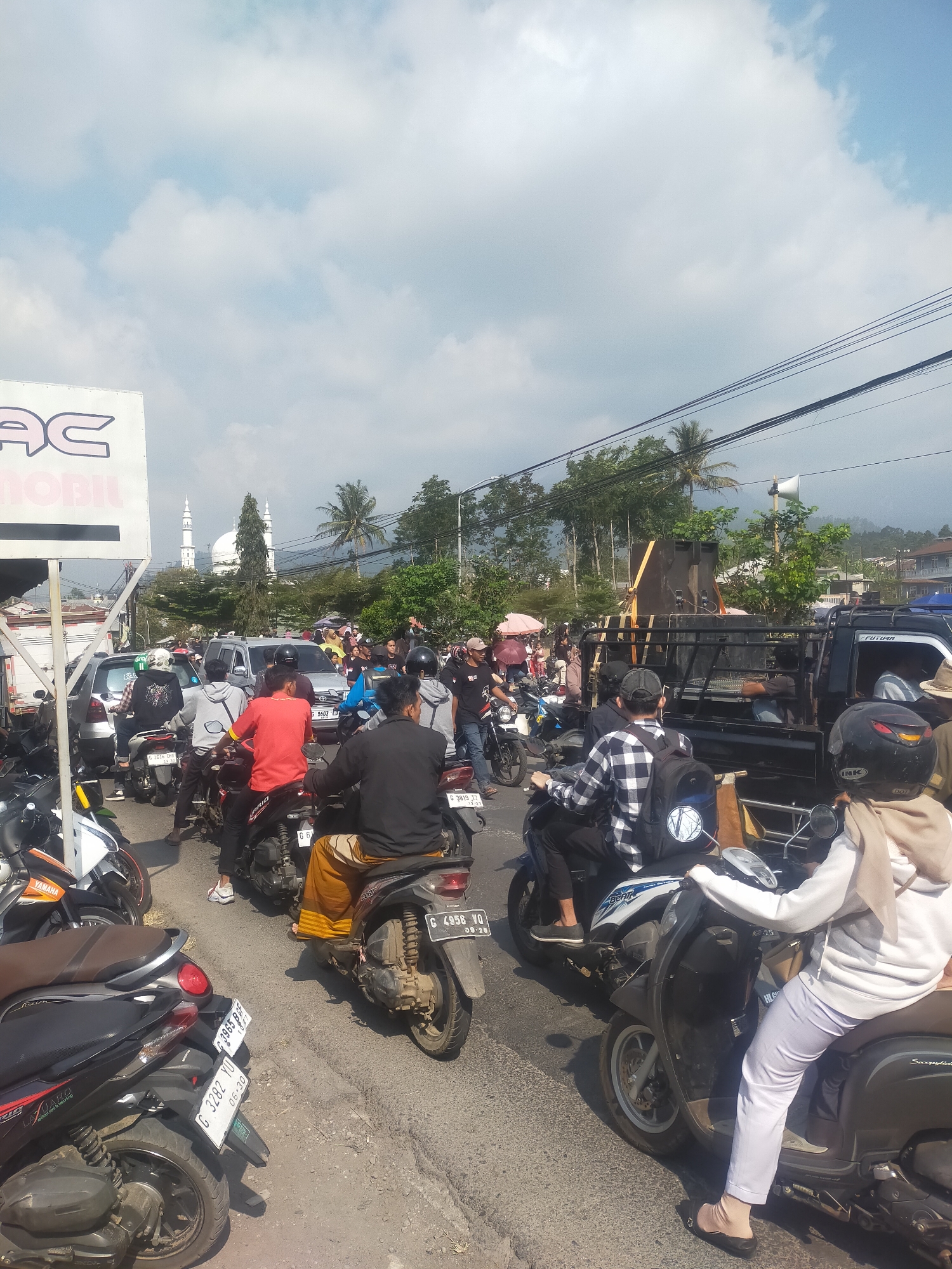 The image depicts a traffic jam on a street in Tuwel, Indonesia, under a partly cloudy sky. Numerous motorcycles and cars are visible, most of them stopped. People are riding on the motorcycles, some looking ahead, others seemingly waiting. In the background, a white mosque with two minarets is prominent, surrounded by trees and mountains. On the right side of the frame, a black pickup truck is loaded with speakers and sound equipment. A large white sign with the text "AC MOBIL" is positioned on the left side of the image. The road appears to be paved.