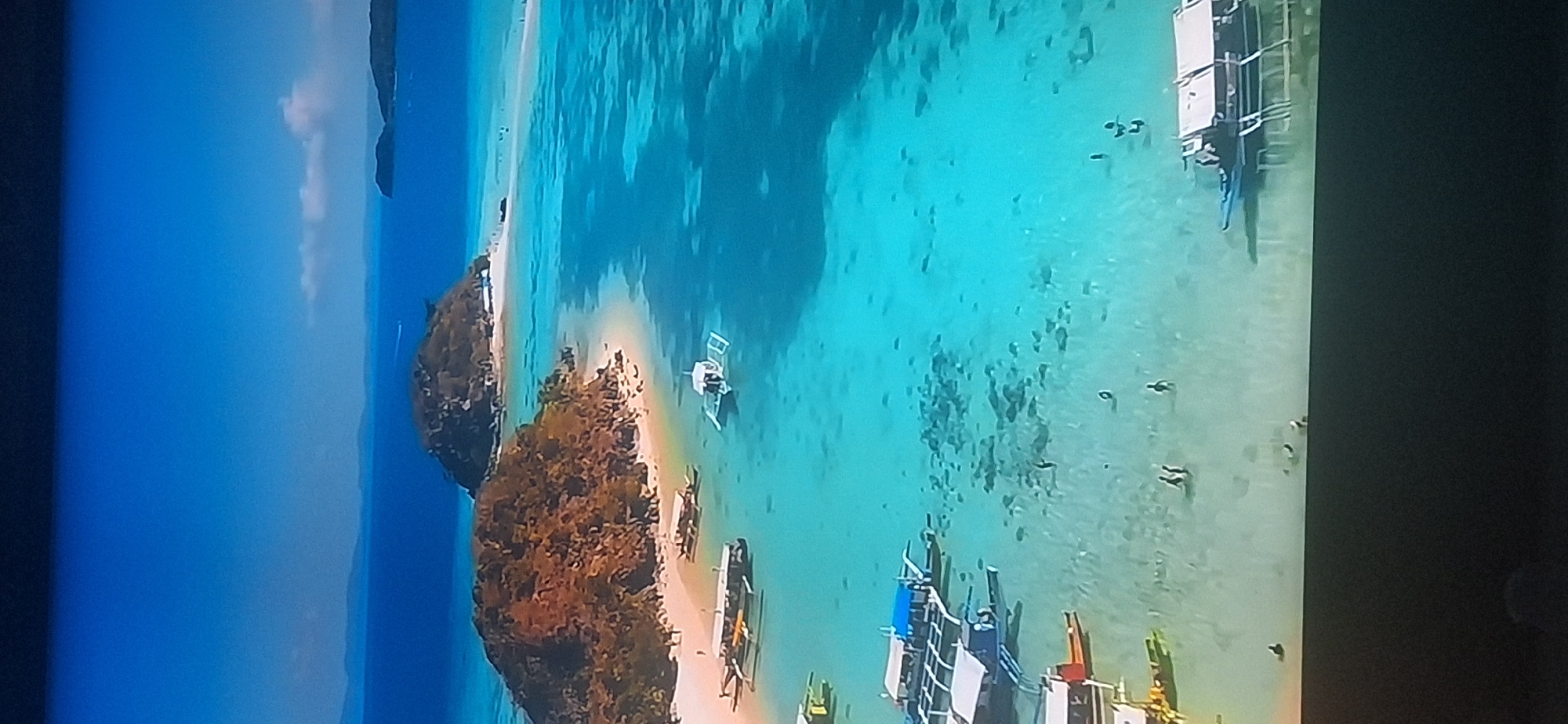 An aerial view shows a tropical paradise with crystal-clear turquoise water, sandy beaches, and lush green islands. Numerous small boats, some with thatched roofs, are scattered across the shallow water near the shore. Several people are visible wading in the water and walking on the beach. The scene evokes a sense of tranquility and vacation. There is no indication of the city of Bila Tserkva, Ukraine, in this image.