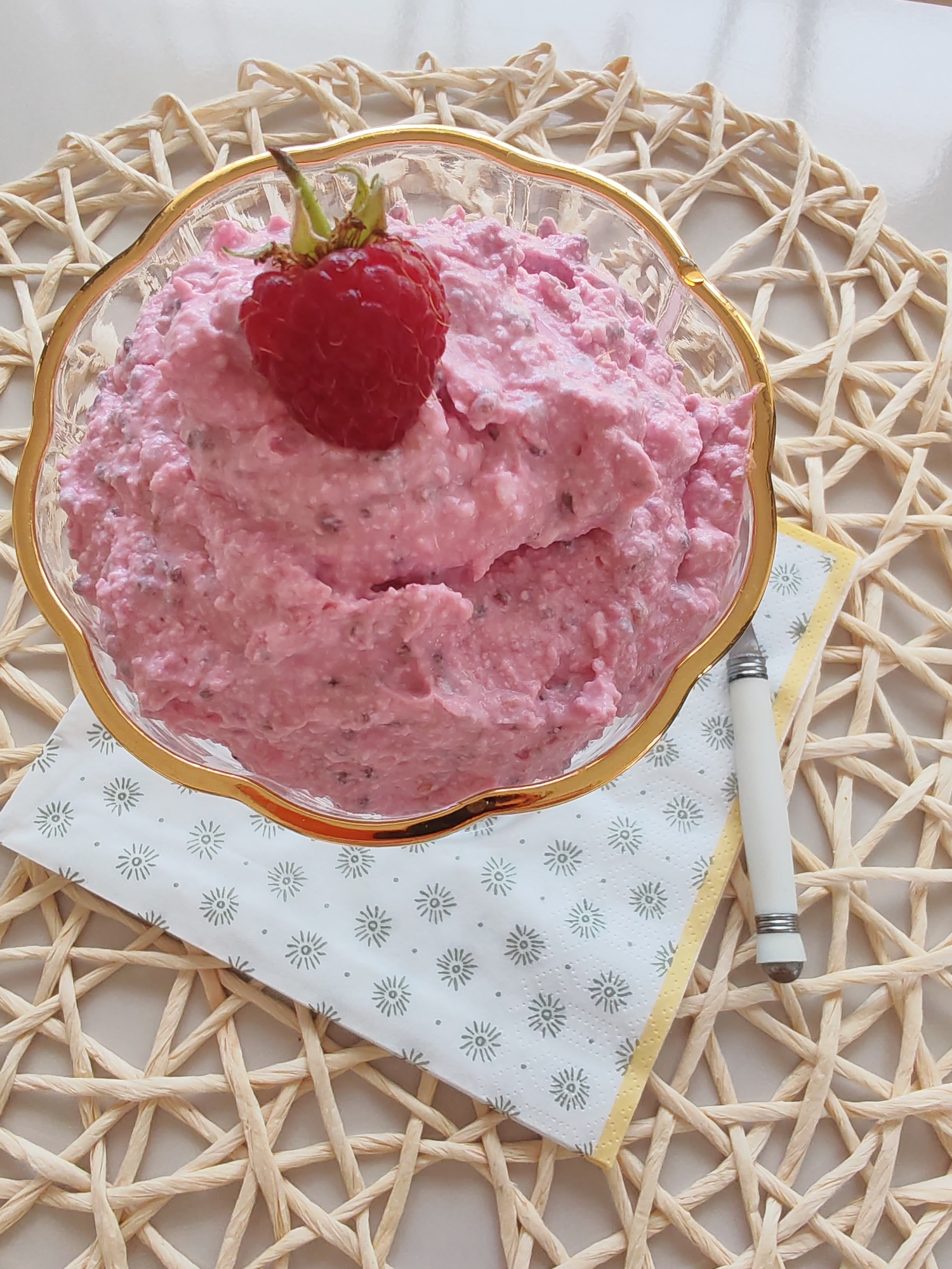 A close-up, top-down shot shows a glass bowl filled with a creamy pink dessert, possibly a mousse or parfait, topped with a single fresh raspberry. The bowl has a decorative gold rim and sits on a woven straw placemat. A white napkin with a delicate sunburst pattern and a yellow trim is placed beneath the bowl and to its right. A small white spoon with silver accents rests on the napkin. The lighting is soft, casting gentle shadows. The image is indoors, on a neutral-colored surface. There are no people or animals present. The location is not identifiable within the city of Sabunchu Raion, Azerbaijan, or any other specific geographic location, as the image does not contain any discernible landmarks or contextual clues.