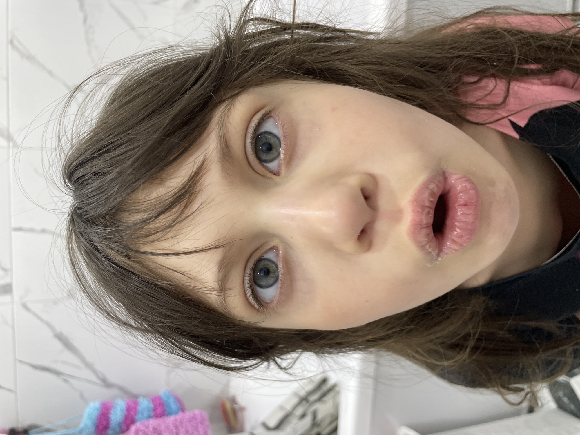 The image presents a close-up portrait of a young individual, likely a child, with fair skin, long, slightly disheveled dark brown hair, and striking light blue-grey eyes. The subject is looking directly at the camera with wide, alert eyes and an open mouth forming an 'o' shape, conveying an expression of surprise, wonder, or curiosity.
The photo is taken from a low angle, and the subject's head is tilted, causing the entire image to appear rotated. A notable detail is the texture of the subject's lips, which appear chapped or dry, with some light, possibly dried, residue visible around the edges. Long, dark eyelashes frame the prominent eyes.
Parts of the subject's clothing are visible on the right side of the frame, including a pink garment worn over or alongside a black one. The background suggests an indoor setting, likely a bathroom, characterized by a white tiled wall with subtle grey marble-like veining. In the lower-left corner, a soft, textured object with blue and pink stripes, possibly a knitted washcloth or decorative item, is partially visible. Hints of a white fixture, perhaps a sink or counter, are also present in the lower right.
The image captures a candid moment, intimately focusing on the subject's face and expression, with no specific activity beyond being photographed. The bright, even lighting indicates an indoor setting during the daytime or a well-lit interior. No visible text is present in the image. The location context provided is Izhevsk, Russia.