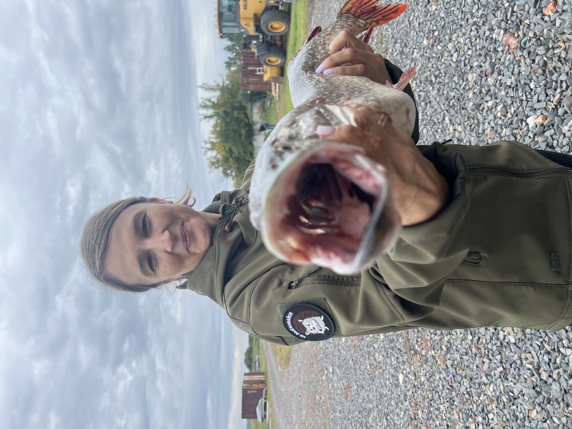 A woman, likely in her 30s or 40s, stands outdoors, holding a large fish with its mouth agape towards the camera. She is wearing a dark green jacket with a patch on the left shoulder that features a hedgehog and Cyrillic text. Her expression is a slight smile, and she is looking directly at the camera. The fish appears to be a pike, with a long body, sharp teeth, and reddish fins. The background suggests an outdoor industrial or rural area. To the left, there are gravel paths and what appear to be shipping containers or sheds. In the mid-ground and to the right, a yellow construction vehicle, possibly a loader, is visible. The sky is overcast with clouds. The overall scene appears to be in Ново-Пичуго, Russia, given the context of the original image's metadata.