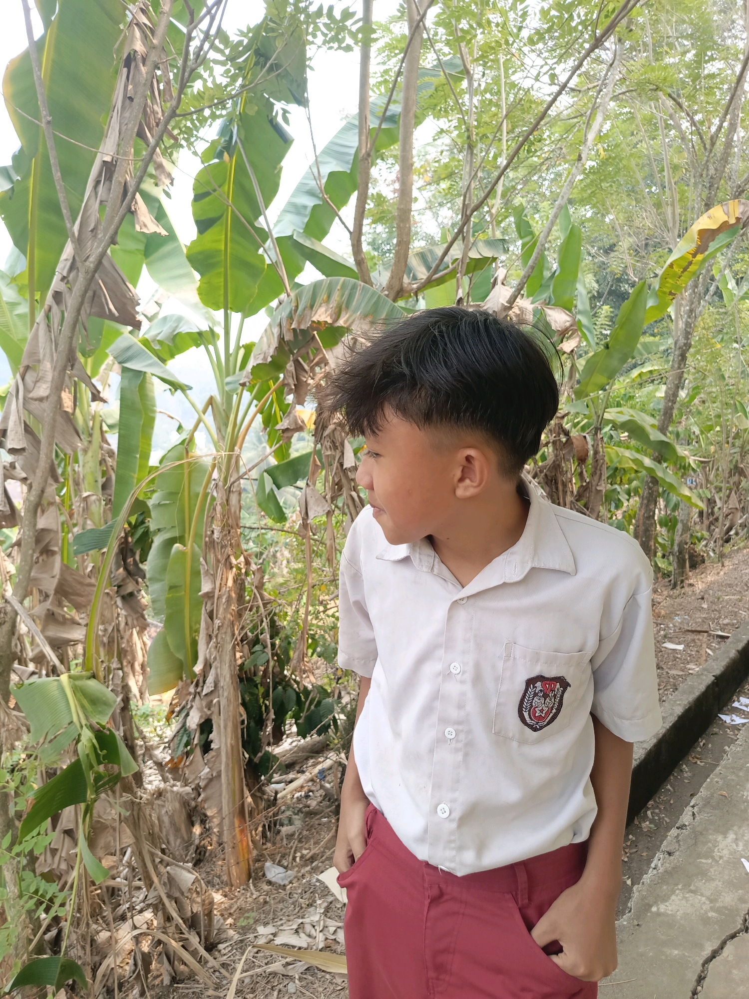 The image depicts a young boy in a light-colored collared shirt and dark red pants, standing outdoors amidst lush vegetation. He is looking to his left, with his hands in his pants pockets. The background is filled with large green leaves, likely from banana trees, and tall, thin trunks, some appearing dry and fibrous. The ground is covered with dry leaves, dirt, and some scattered debris. To the right of the boy, a concrete structure, possibly a drainage ditch or curb, runs along the edge of a path. The overall environment suggests a rural or semi-rural setting. The boy's school uniform indicates he may be a student. The location is Taba Tinggi, Indonesia.