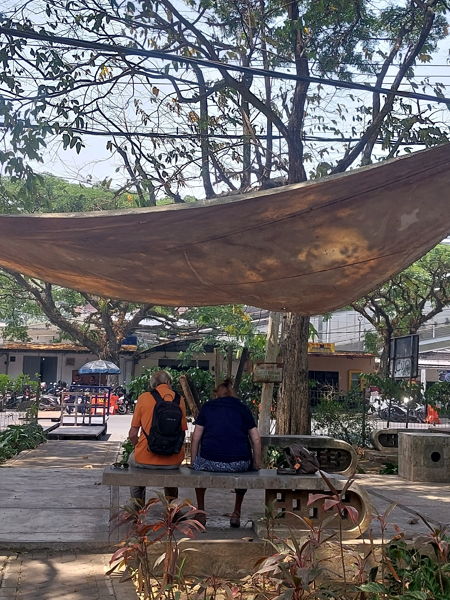 The image depicts an outdoor scene in Malang, Indonesia, under a large, brown, textured canopy stretched between trees and poles. Two individuals are seated on a concrete bench. The person on the left, seen from the back, wears an orange shirt and a black backpack. The person on the right, also seen from the back, wears a dark blue shirt and patterned shorts. A dark-colored bag rests on the bench between them. In the background, there are trees with dense foliage, a street with parked motorcycles, buildings with tiled roofs, and a blue umbrella. Sunlight filters through the canopy and tree branches, casting shadows on the ground. A wooden sign with text is visible on a pole to the left. The overall atmosphere suggests a public outdoor space, possibly a park or resting area.